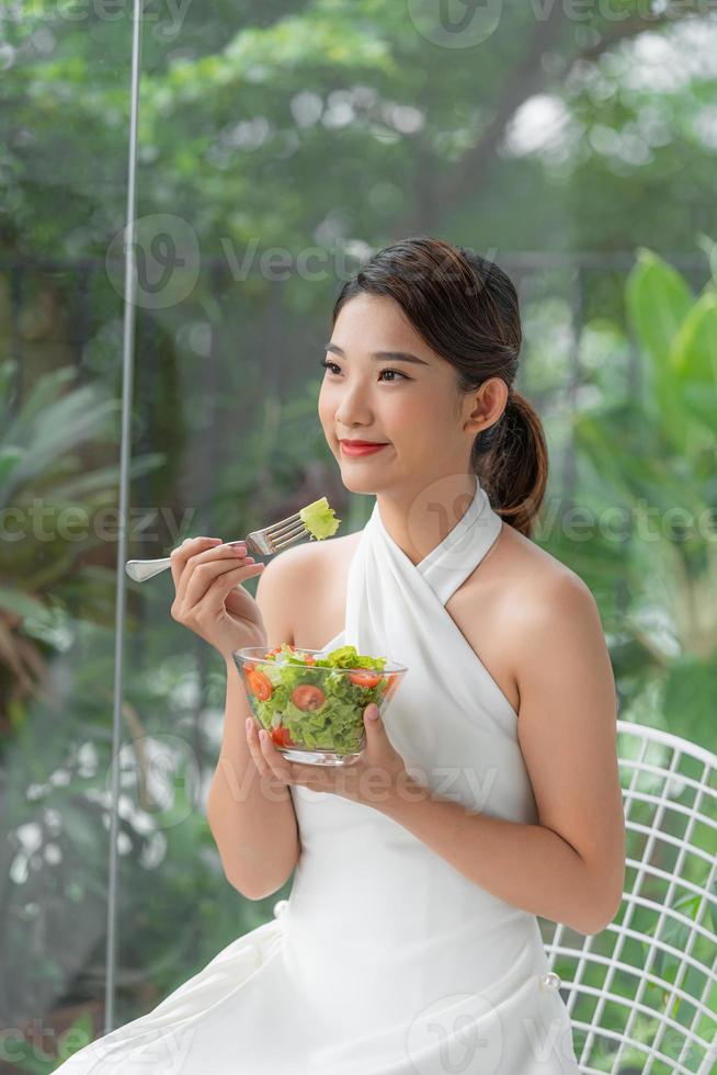 mulher sorridente asiática atraente isolada na sala de estar em casa comendo salada foto