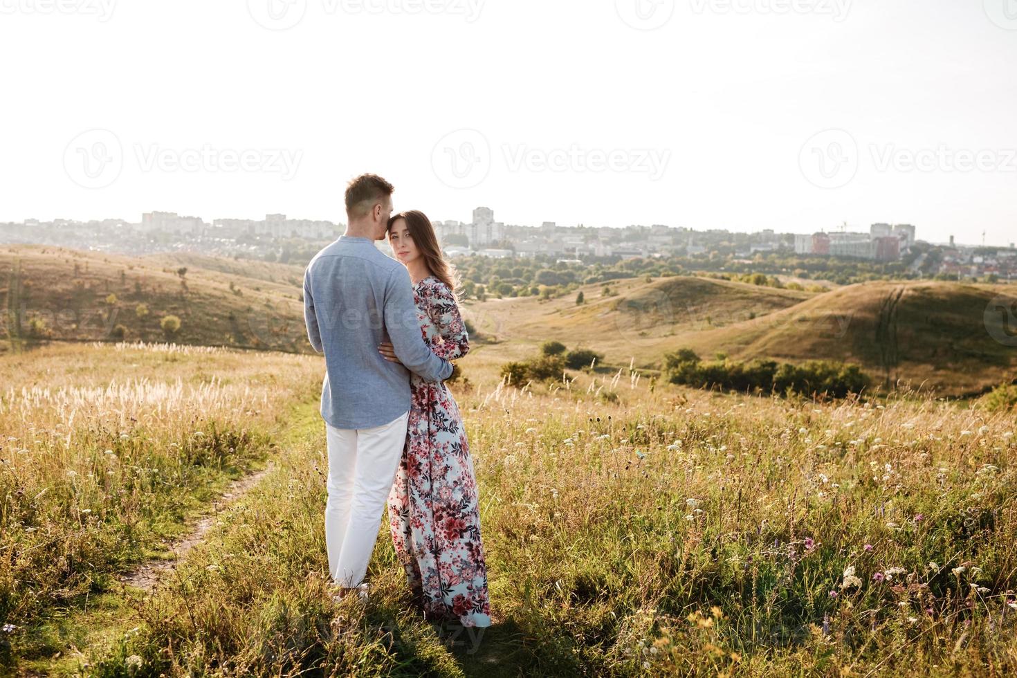 lindo casal jovem abraçando em um campo com grama ao pôr do sol. homem elegante e mulher se divertindo ao ar livre. conceito de família. copie o espaço. foto