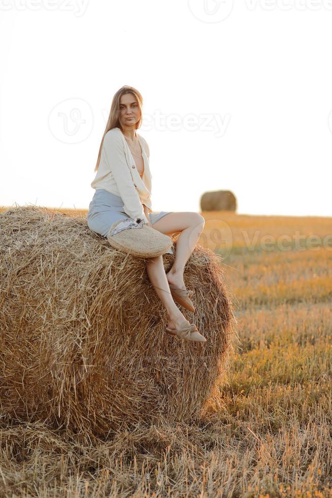 jovem mulher com cabelo comprido, vestindo saia jeans, camisa leve e saco de palha na mão, sentado no fardo no campo no verão. retrato feminino na cena rural natural. conceito de ecoturismo ambiental. foto