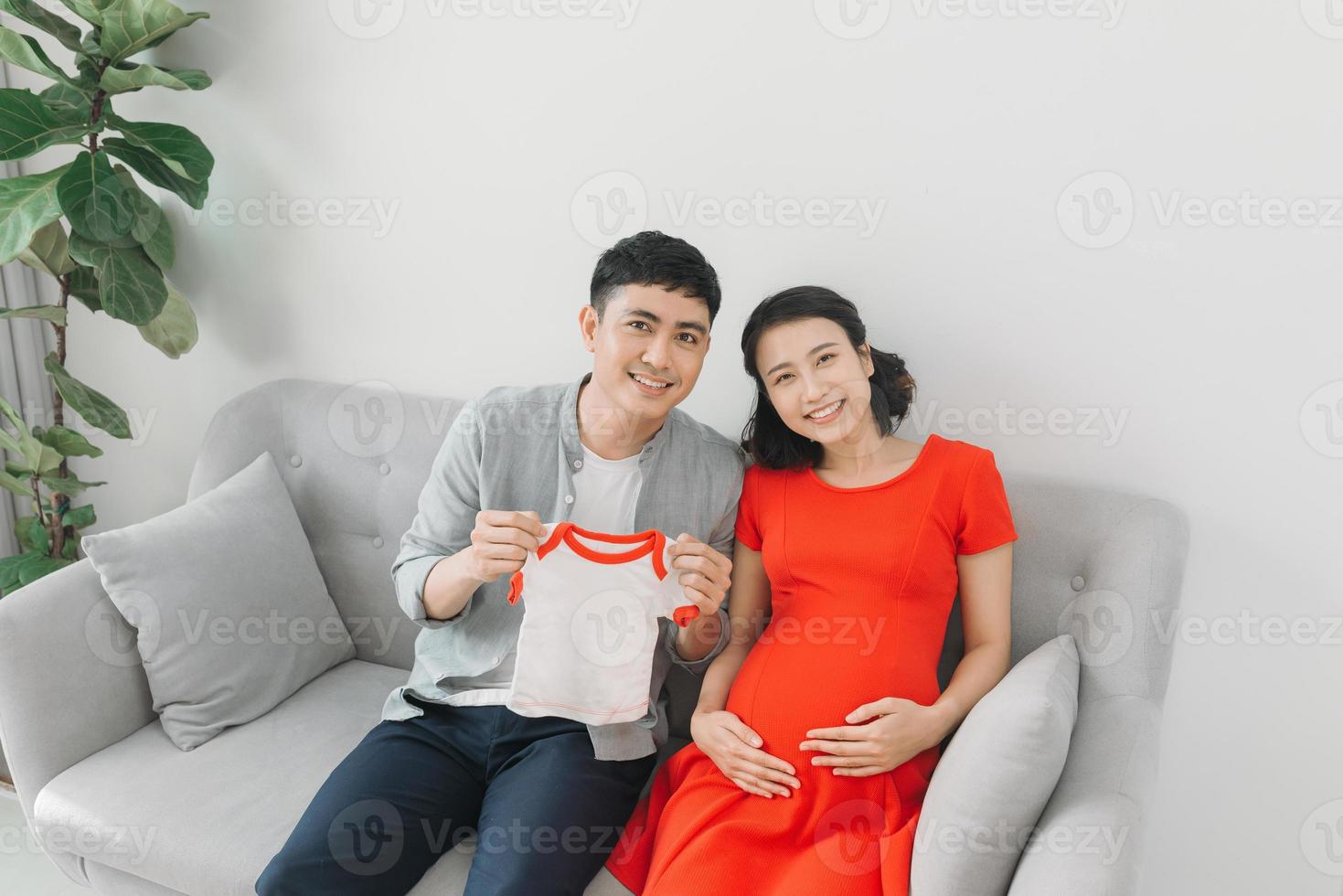 retrato de uma mulher grávida feliz segurando a camiseta do bebê e do marido em um sofá em casa foto