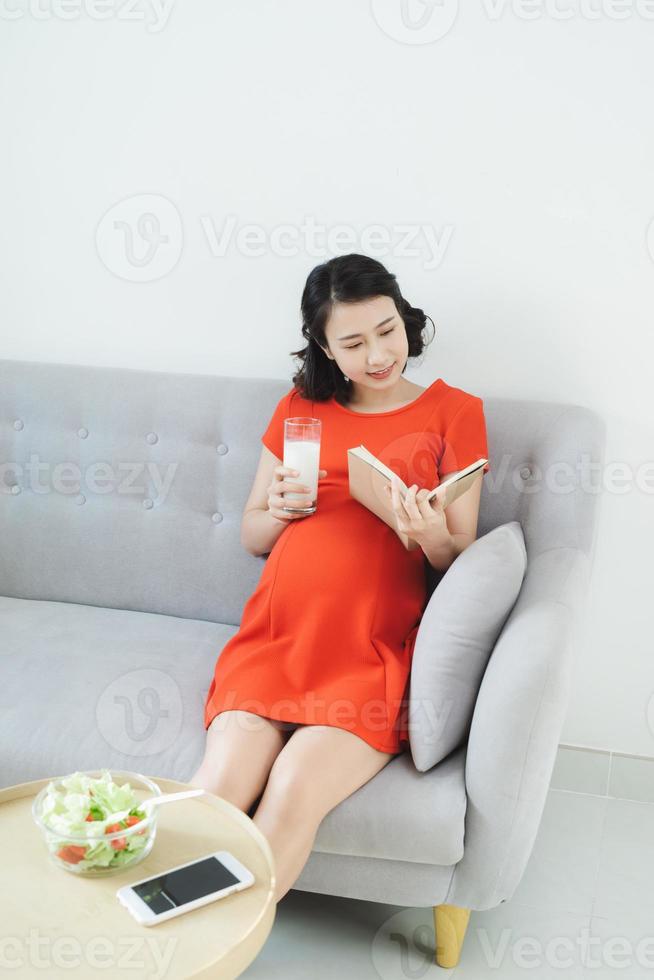 jovem mulher asiática lendo livro relaxando no sofá e bebendo leite. foto