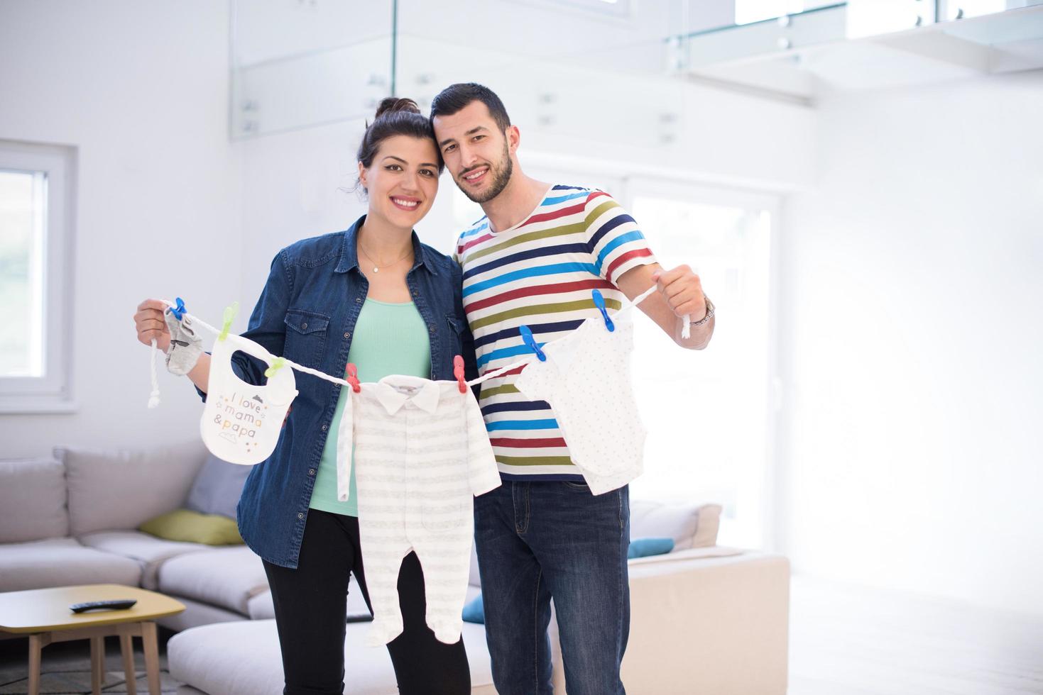 jovem casal segurando roupas de bebê em casa foto