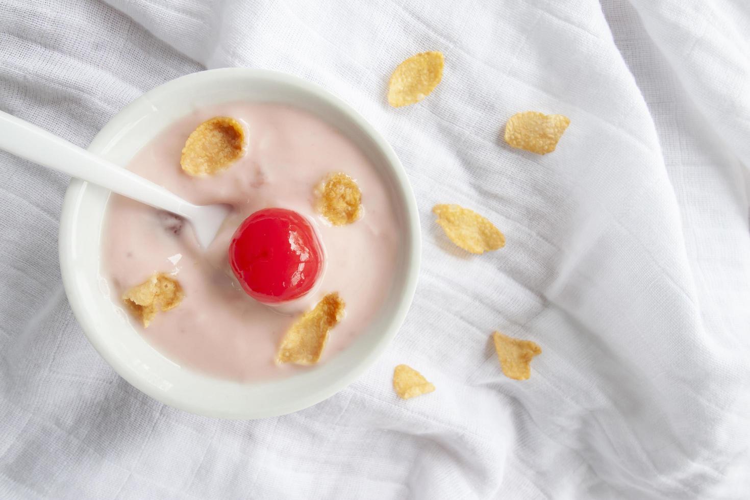 iogurte, cereja vermelha, milho seco e colher em tigela branca sobre fundo de toalha de mesa branca. foto