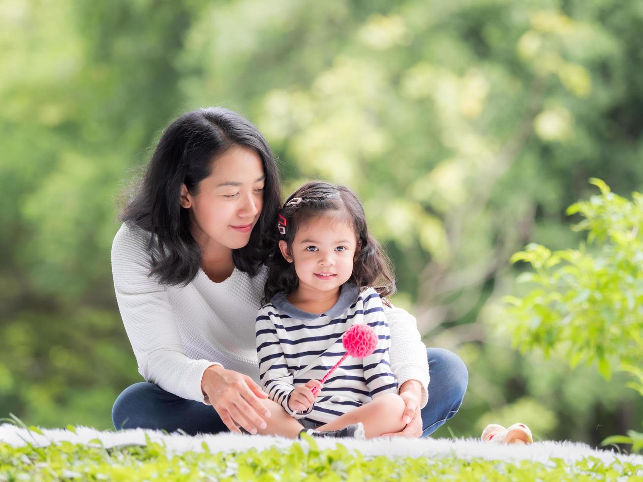 menina asiática sentada com a mãe no pano, relaxando e aprendendo fora da escola para desfrutar no parque natural foto