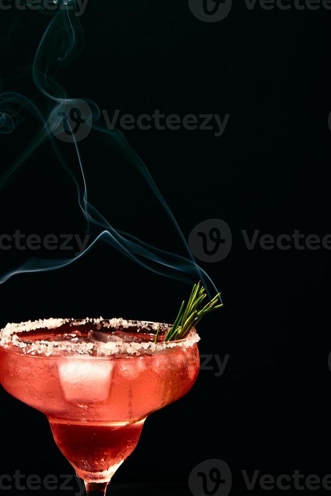 cocktail alcoólico ou não alcoólico do tipo margarita com gelo e ramo de alecrim fumegante em fundo escuro foto