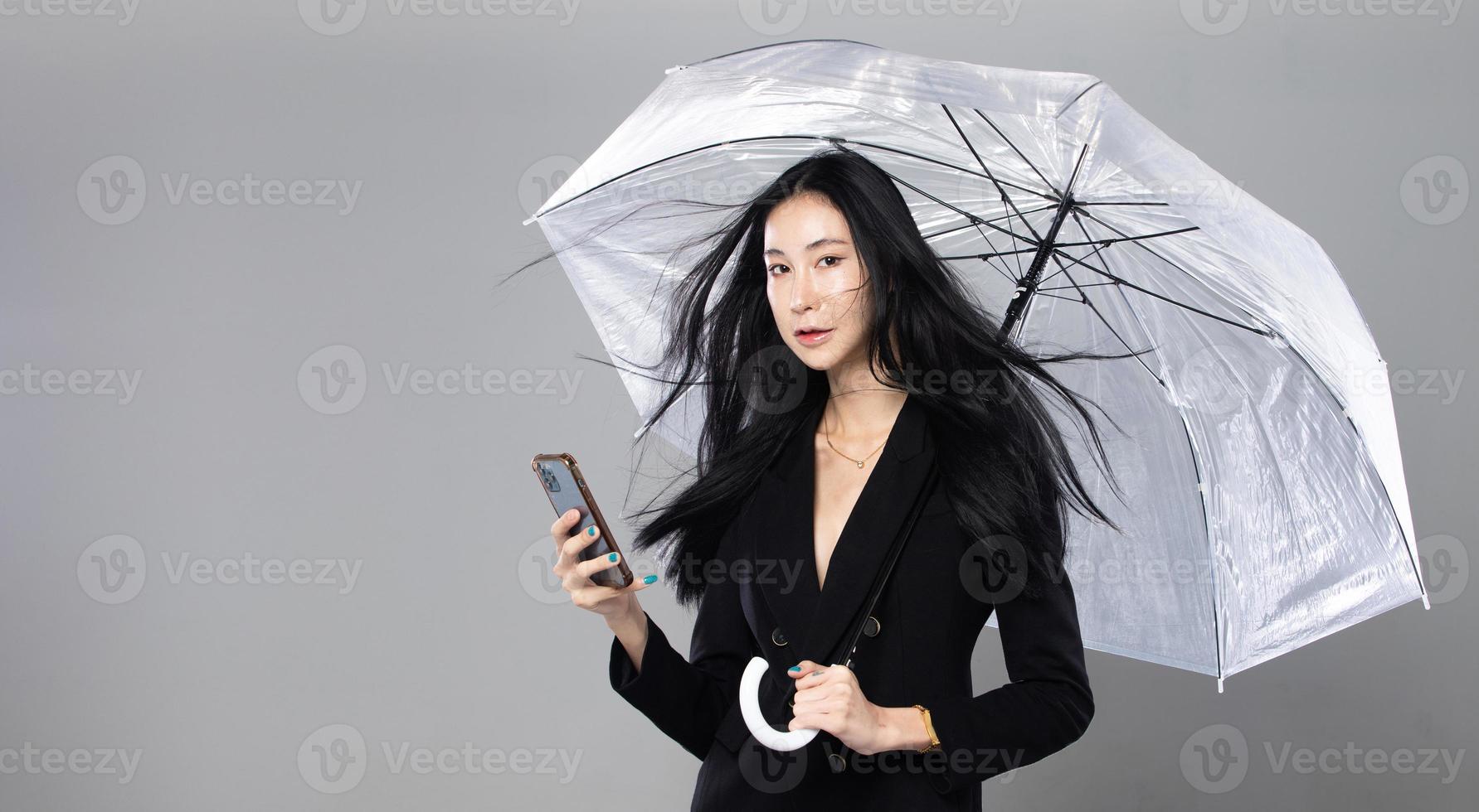 mulher transgênero asiática com longos cabelos lisos pretos, golpe de vento no ar. fêmea segura telefone e guarda-chuva contra tempestade de vento, sentindo moda sensual sexy, espaço de cópia isolado de fundo cinza foto