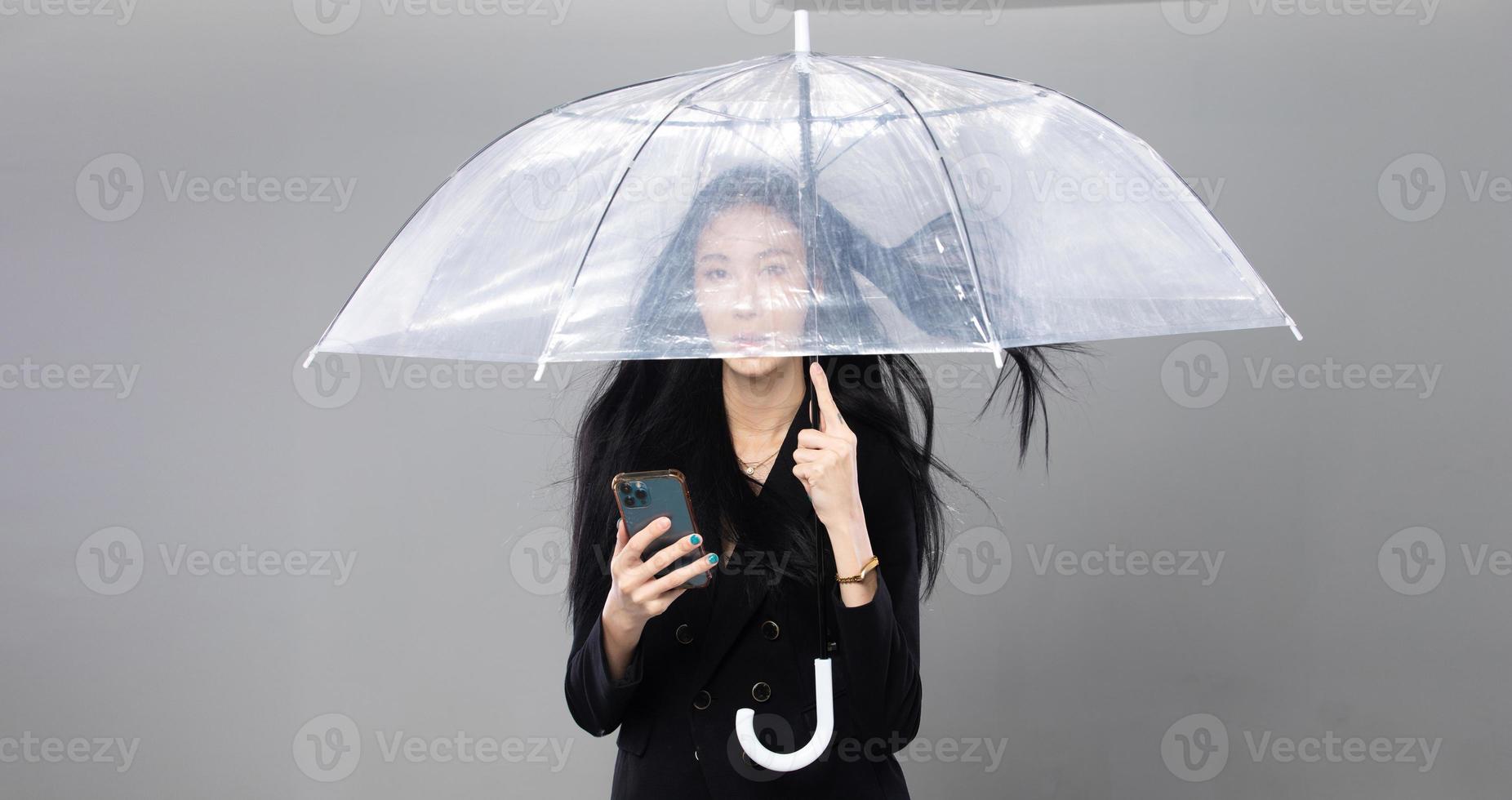 mulher transgênero asiática com longos cabelos lisos pretos, golpe de vento no ar. fêmea segura telefone e guarda-chuva contra tempestade de vento, sentindo moda sensual sexy, espaço de cópia isolado de fundo cinza foto