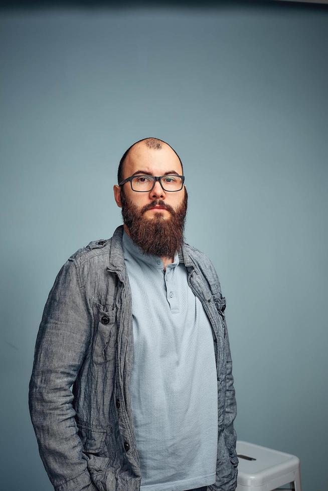 estilo de vida jovem bem sucedido com óculos, barba, jaqueta jeans elegante olhando para a frente, retrato masculino no estúdio em um fundo uniforme. foto