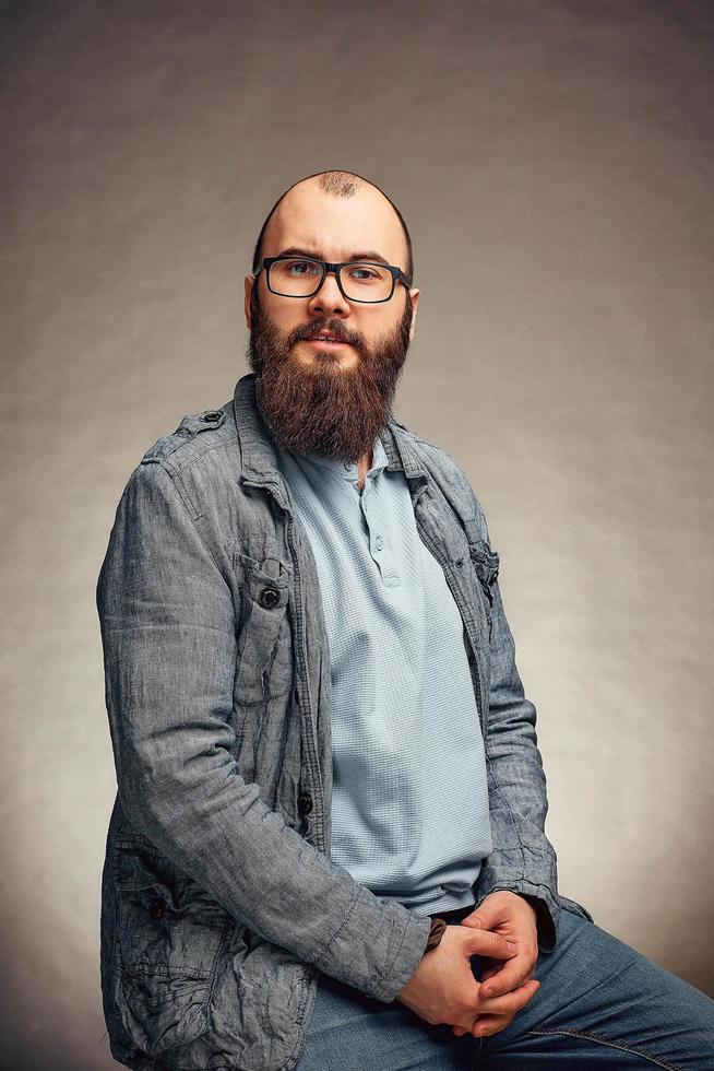 estilo de vida jovem bem sucedido com óculos, barba, jaqueta jeans elegante olhando para a frente, retrato masculino no estúdio em um fundo uniforme. foto