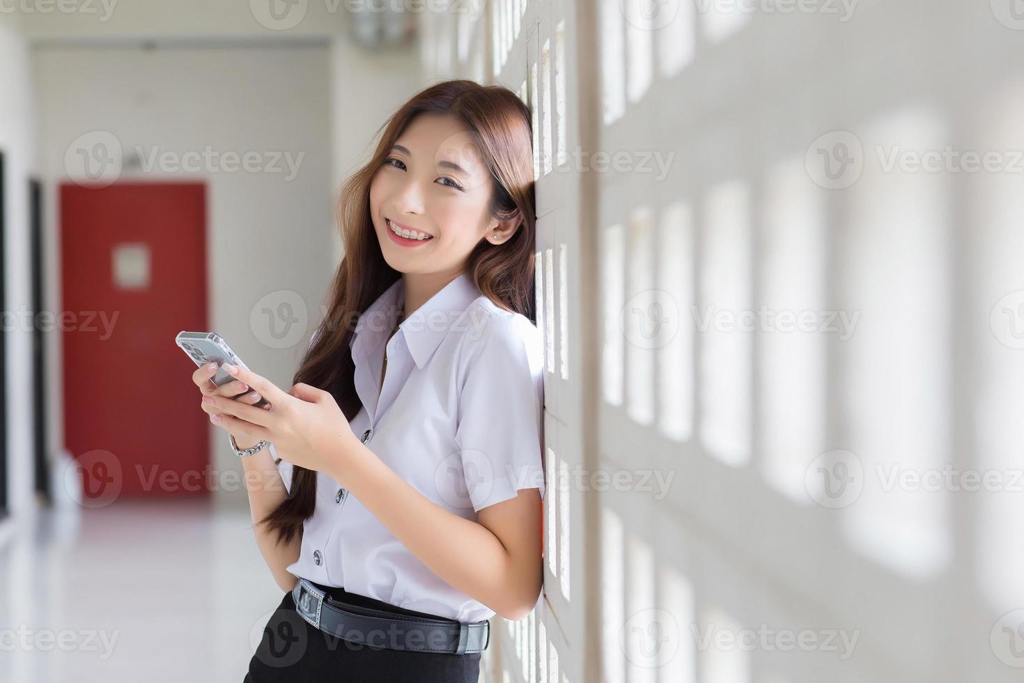 retrato de uma estudante tailandesa asiática de uniforme está de pé sorrindo alegremente e com confiança enquanto estiver usando o smartphone no prédio da universidade com plano de fundo. foto