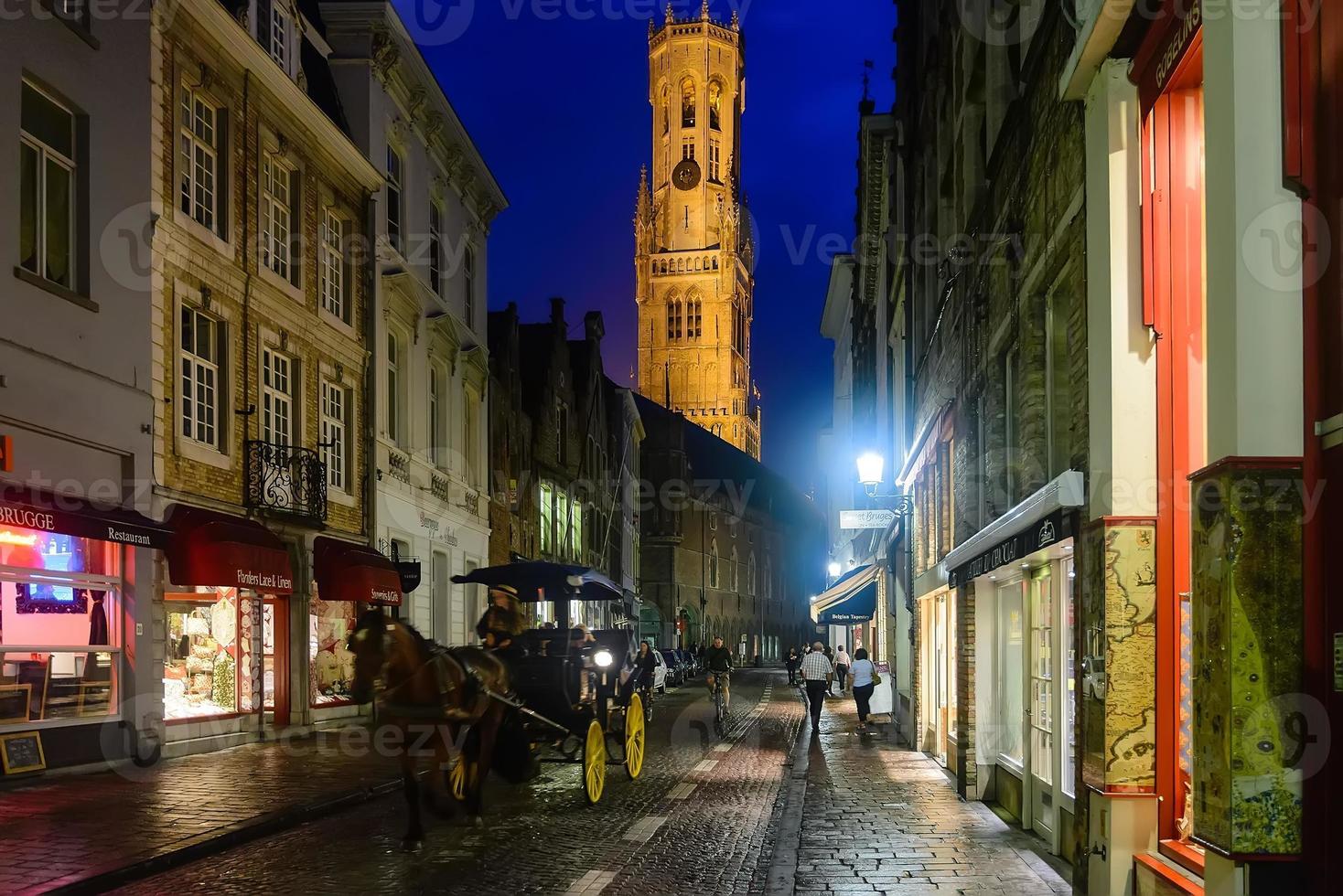 antiga rua estreita pavimentada na cidade medieval de bruges por luzes noturnas refletidas em paralelepípedos. vista para antigas casas mercantes, befry, treinador e pessoas andando de bicicleta. foto