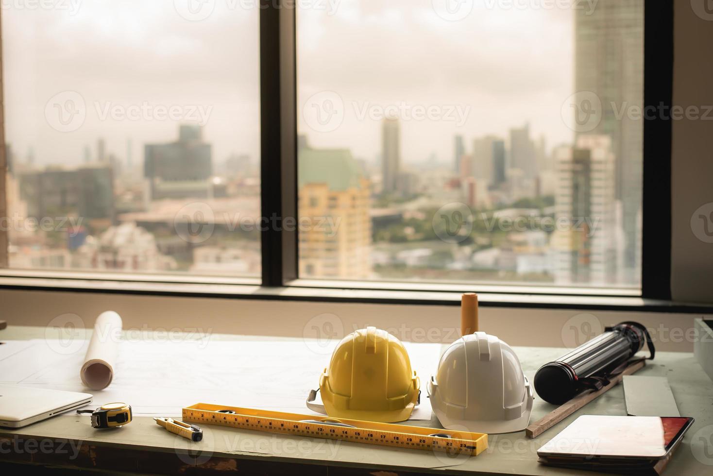 capacete, tablet, laptop, rolo de planta ou plano arquitetônico, régua, instrumento de medição na mesa do trabalhador da equipe de construção e um prédio alto ao fundo. foto