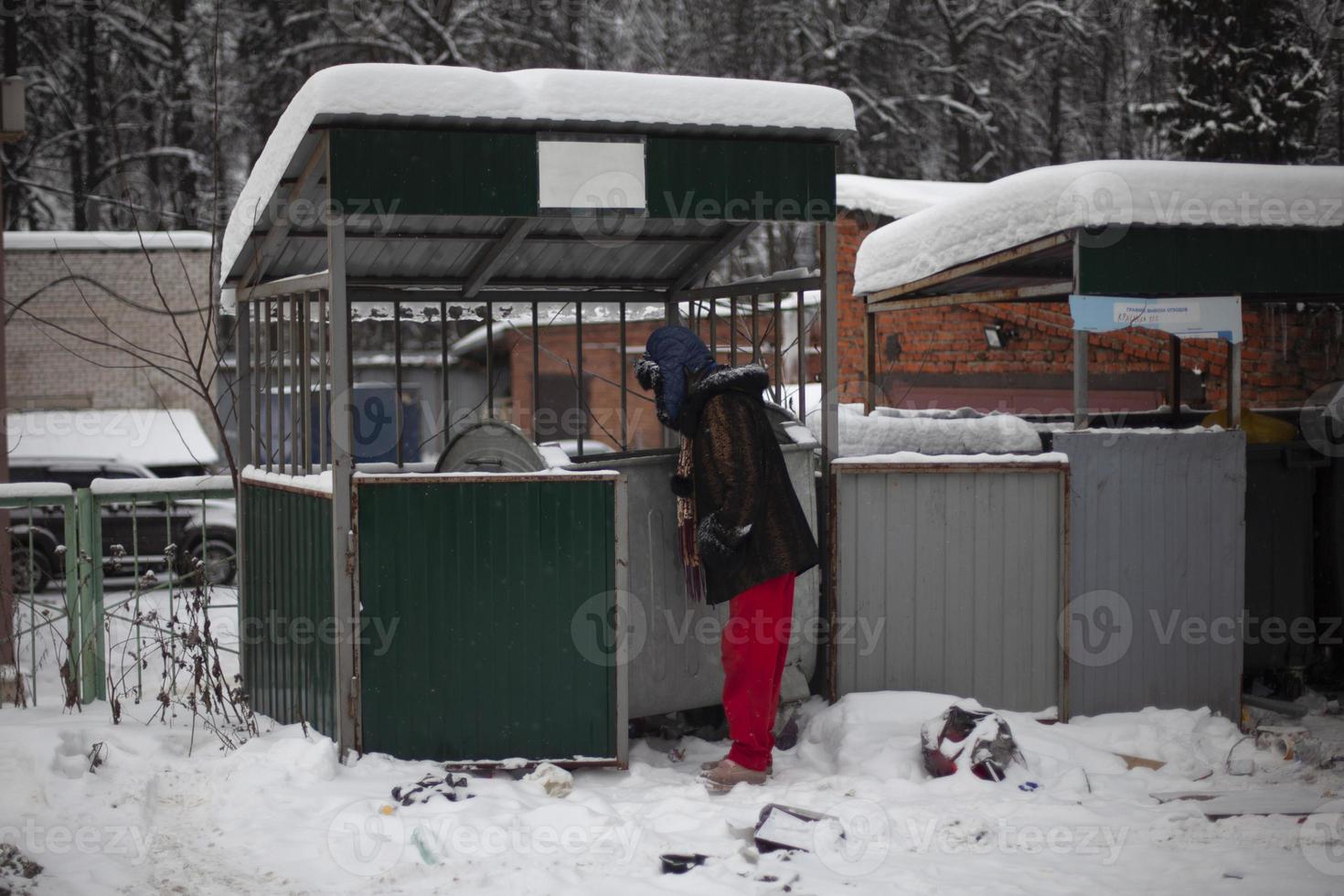 homem está procurando comida no lixo. homem olha para a lixeira no inverno. pobre na rua. foto