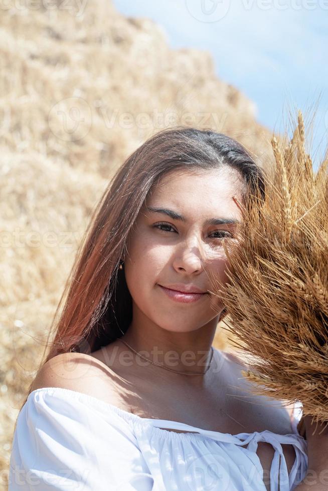 jovem mulher sentada no palheiro no campo colhido segurando o buquê de trigo foto