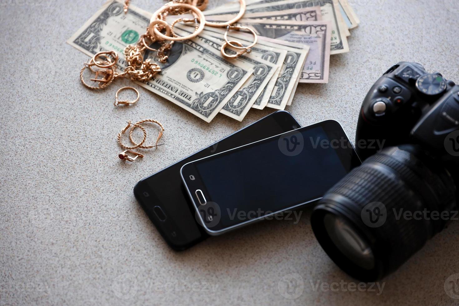 muitos anéis, brincos e colares dourados caros com grande quantidade de notas de dólar perto de smartphones e câmera digital slr. loja de penhores foto