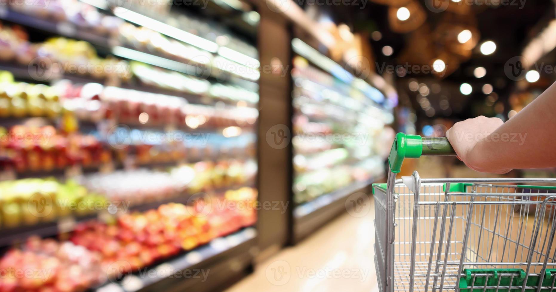mão de mulher segura carrinho de compras de supermercado com borrão abstrato frutas e vegetais orgânicos nas prateleiras na mercearia desfocado fundo claro bokeh foto