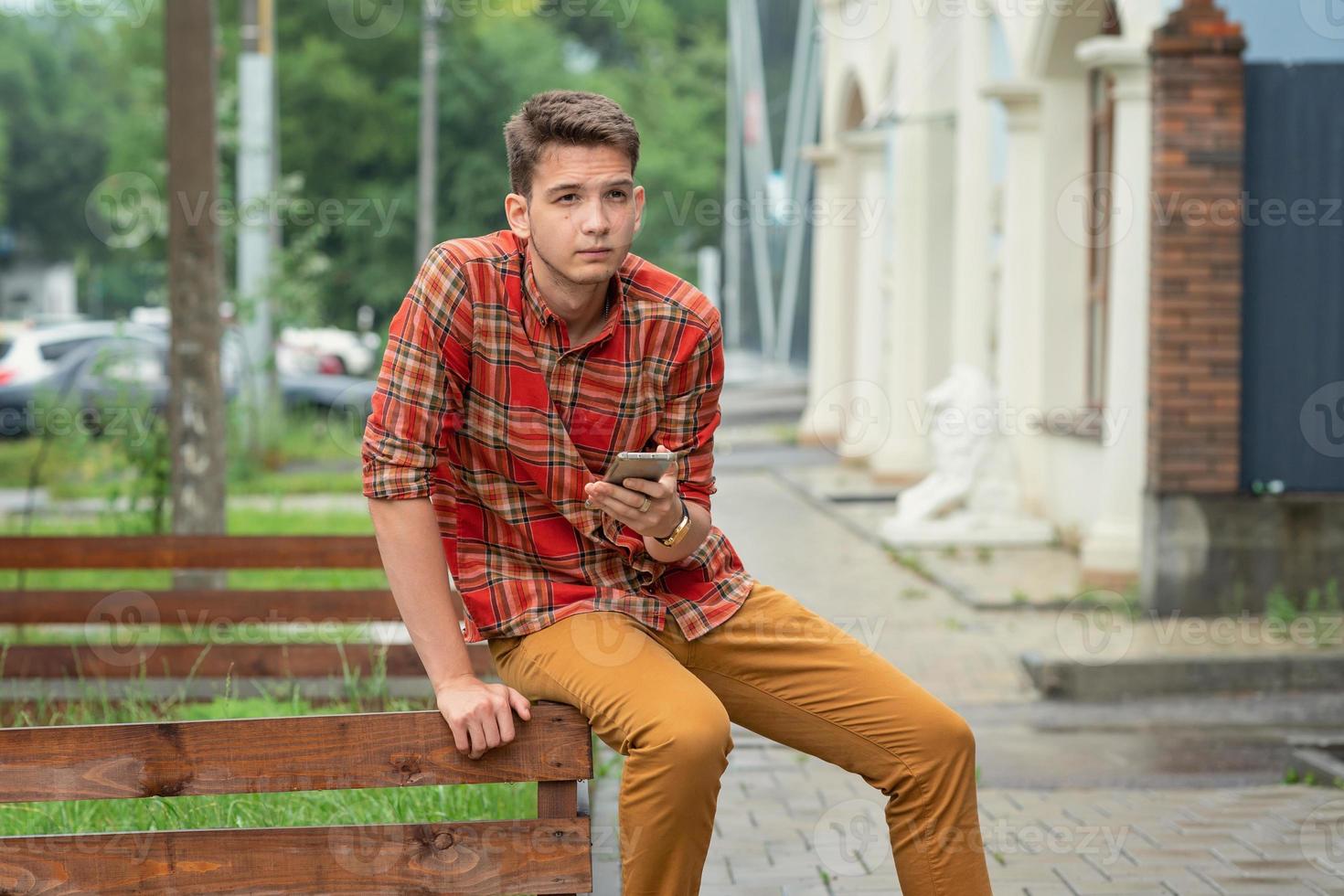 um jovem de camisa xadrez senta-se em uma grade de madeira, segura um telefone nas mãos e digita uma mensagem foto