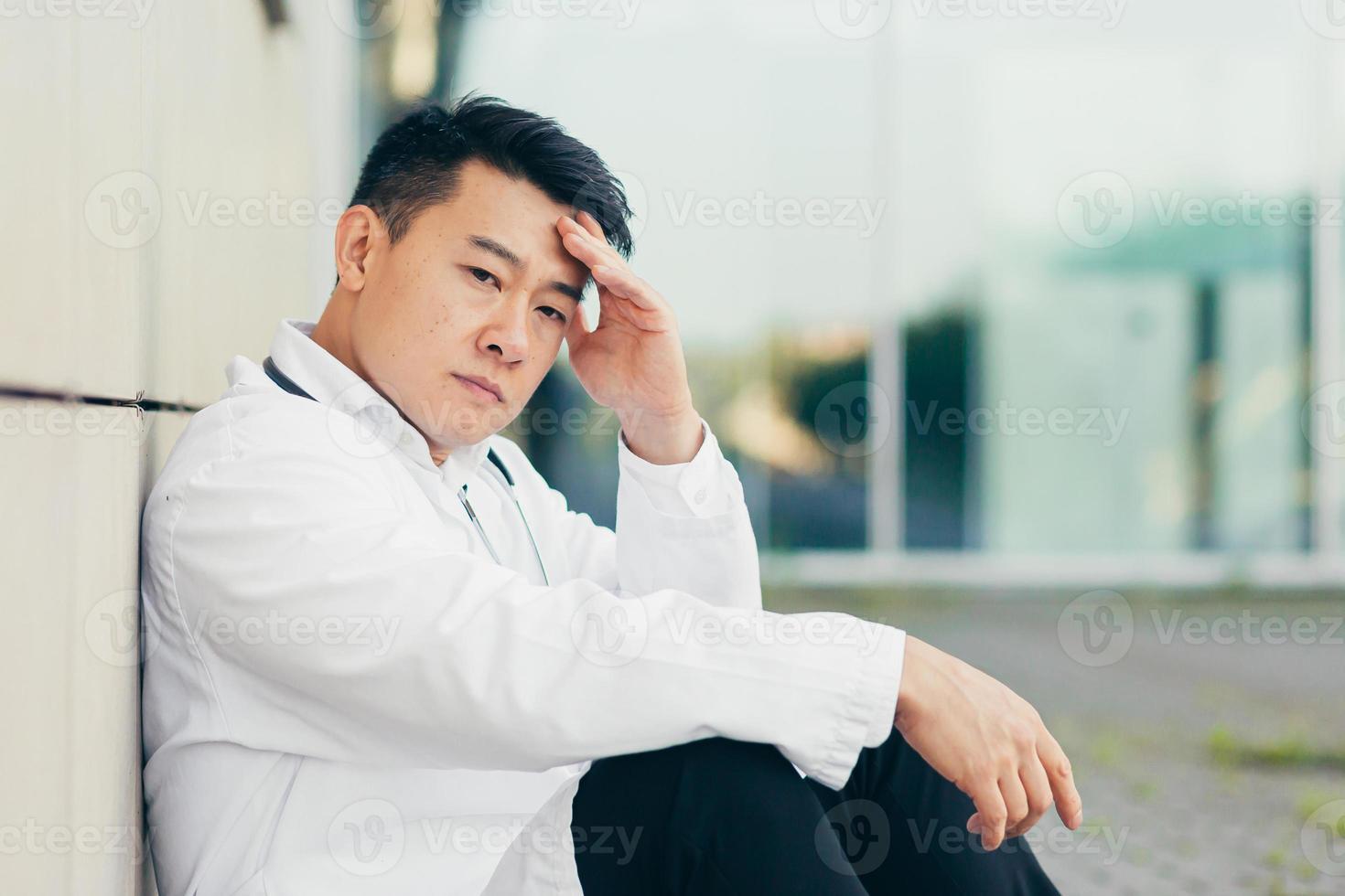 retrato médico homem asiático cansado depois do trabalho sentado no chão perto da clínica desapontado foto