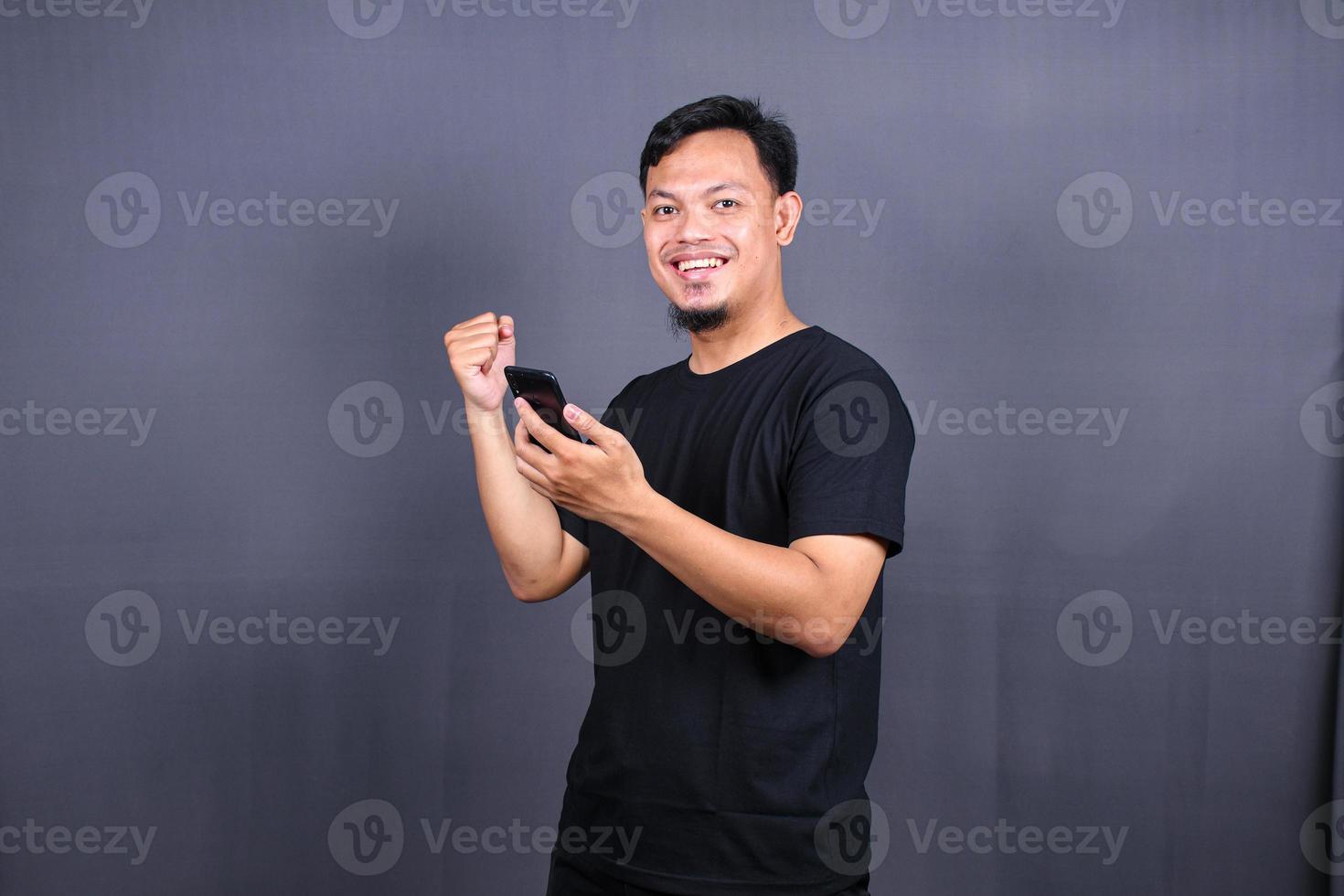 feliz homem asiático segurando um smartphone e ganhando o prêmio. isolado no fundo cinza foto