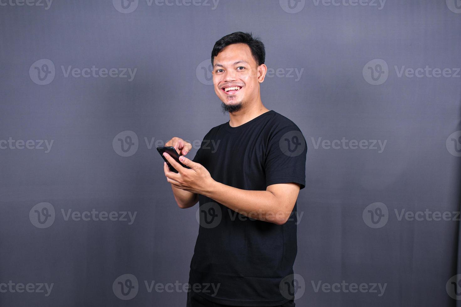 retrato de jovem asiático vestindo camiseta preta em pé isolado sobre fundo cinza, apontando o dedo para o celular foto