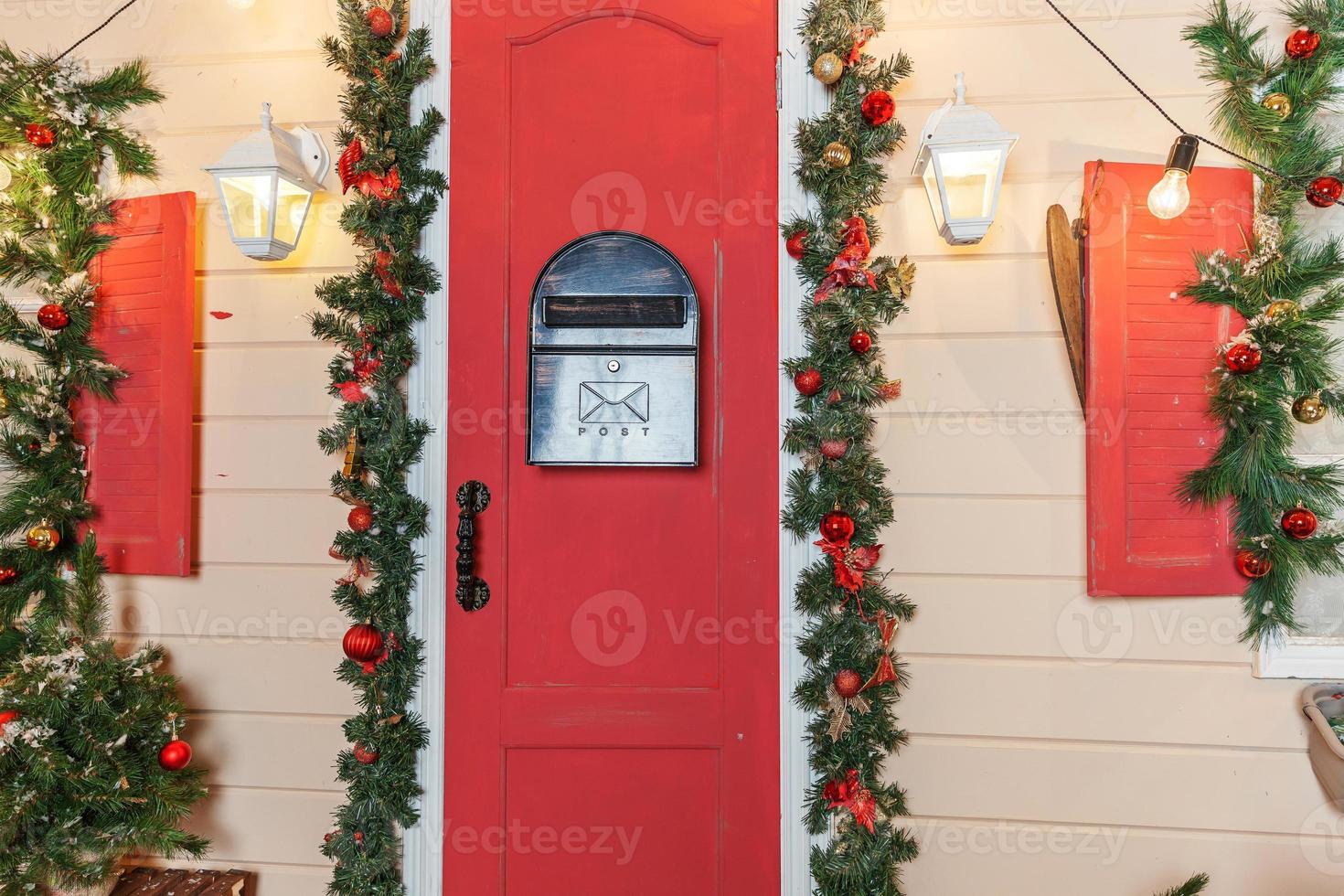ideia de decoração de varanda de natal. entrada da casa com porta vermelha decorada para feriados. guirlanda de grinalda vermelha e verde de galhos de árvores do abeto e luzes na grade. véspera de natal em casa foto