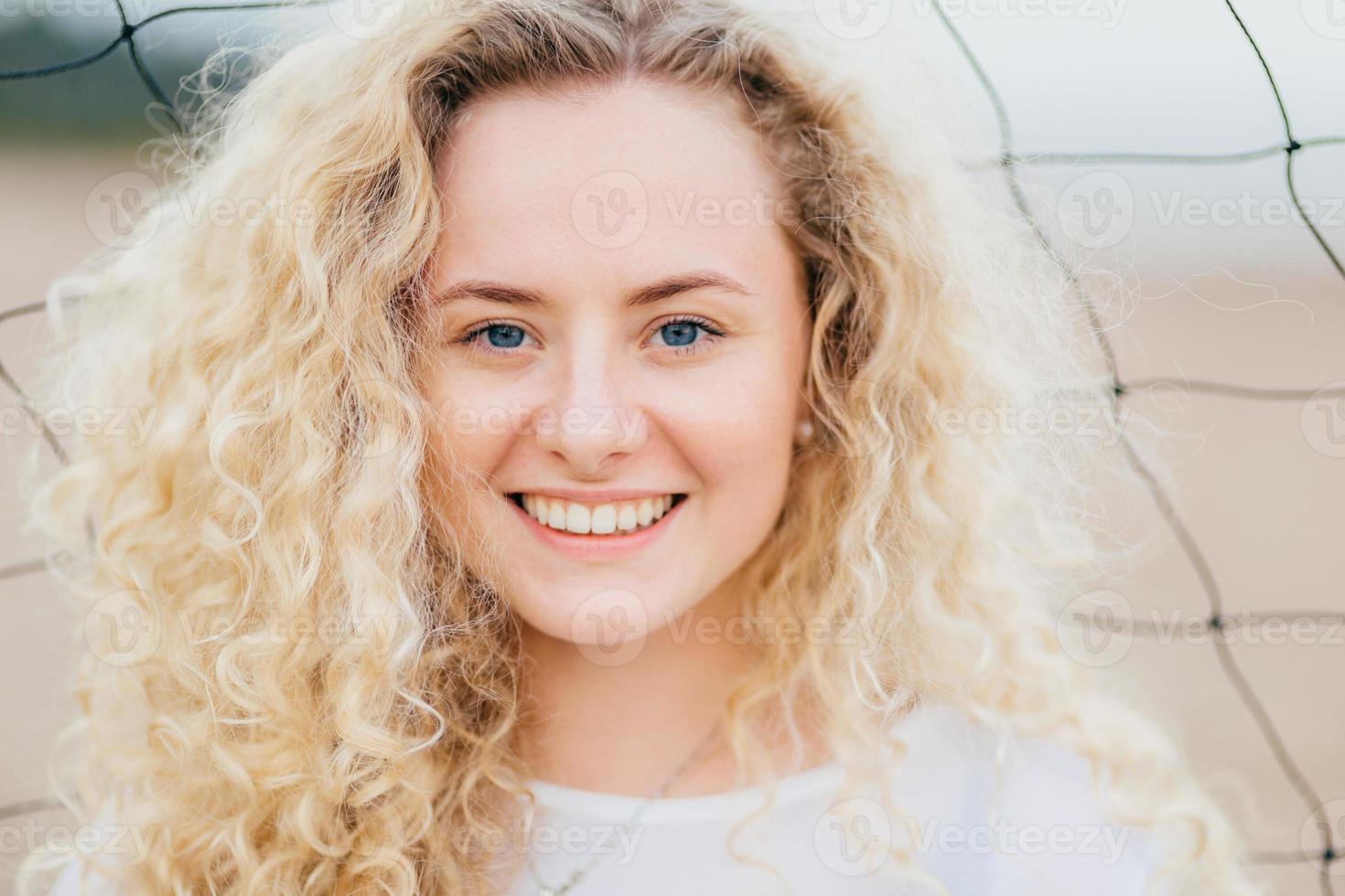 feliz mulher caucasiana encaracolada com sorriso cheio de dentes, tem cabelos claros e encaracolados, olhos azuis, pele limpa, fica ao ar livre, tem uma aparência atraente. close-up retrato de mulher bonita descansa na praia foto