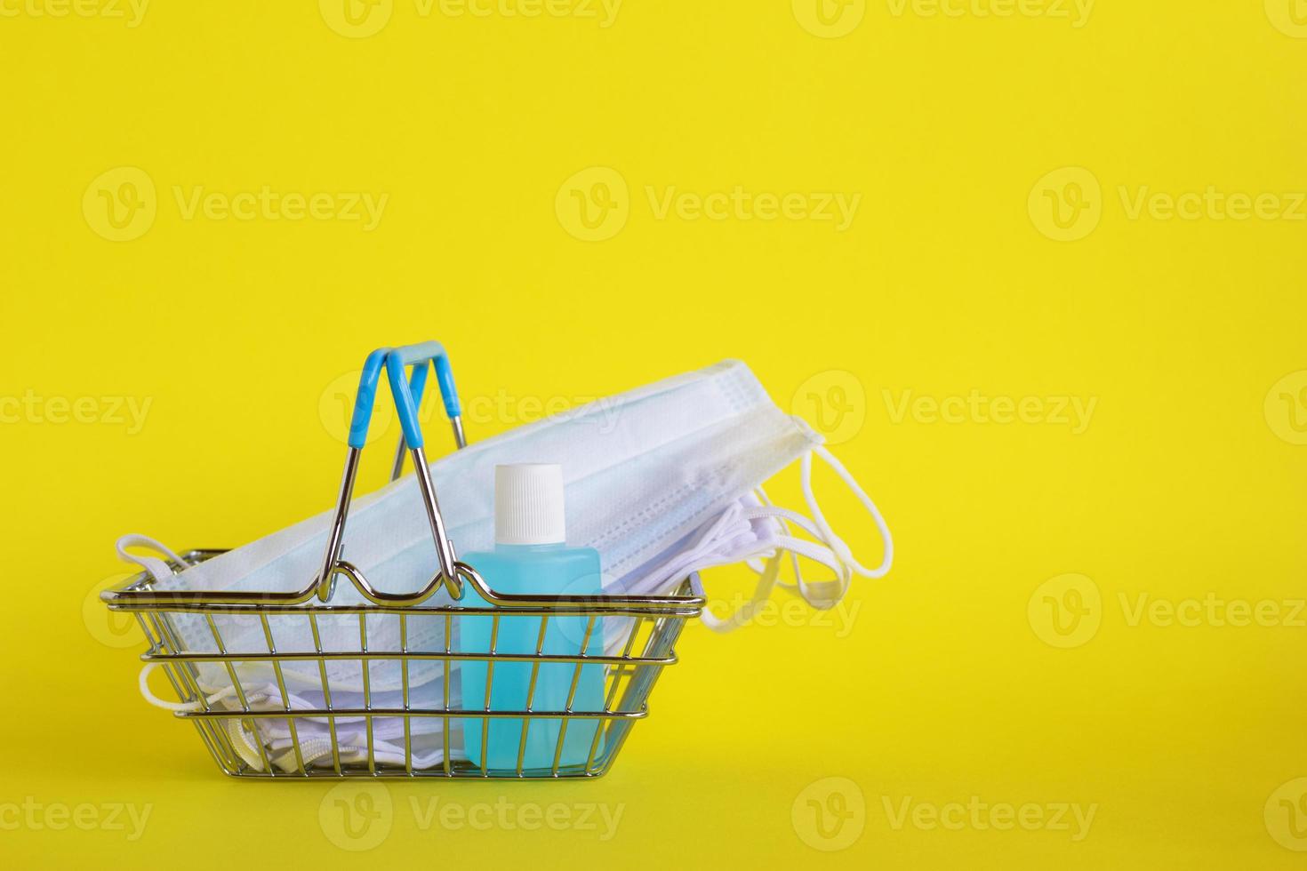 máscaras faciais médicas e garrafa de gel desinfetante para as mãos em uma pequena cesta de compras em fundo amarelo com espaço de cópia. conceito de compras no supermercado no novo estilo de vida normal. foto