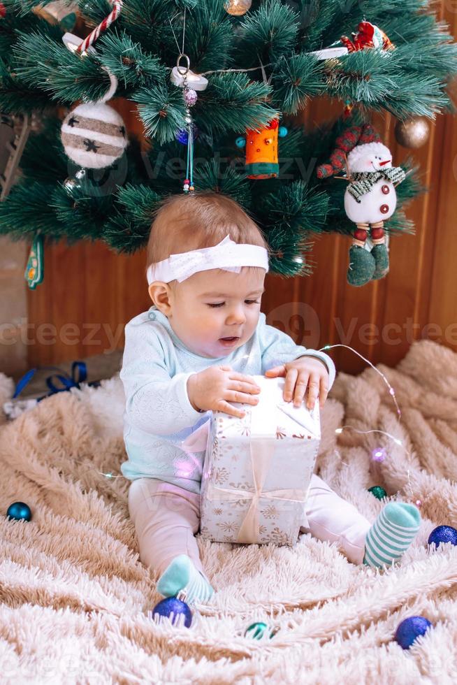 menina bonitinha está sentada perto da árvore de natal em uma manta de pelúcia bege com luzes e decorações de natal e tente abrir o presente de natal. foto