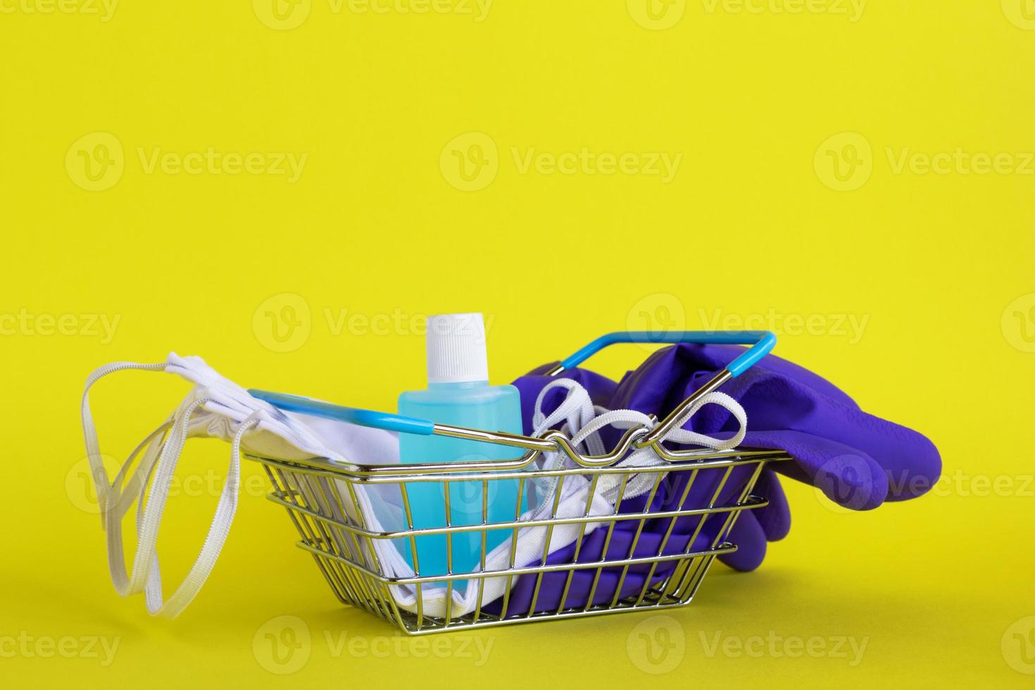 máscaras faciais, par de luvas médicas descartáveis e frasco de gel desinfetante para as mãos em uma pequena cesta de compras em fundo amarelo com espaço de cópia. conceito de compras no supermercado no novo estilo de vida normal. foto