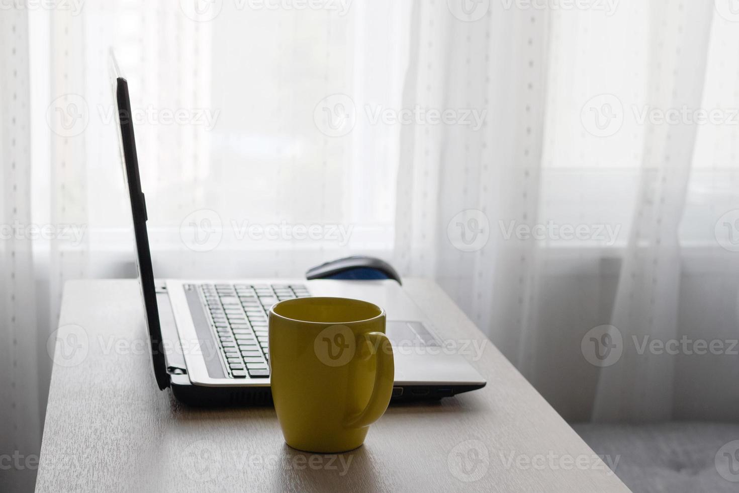 trabalhar em casa na sala de estar. laptop e copo de bebida quente na mesa de madeira perto do sofá e janela. foto
