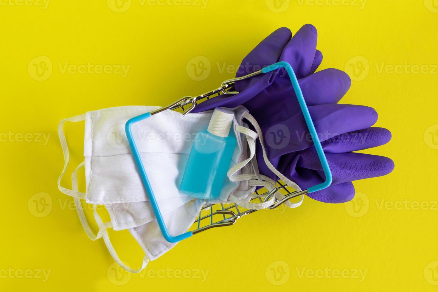 postura plana na cesta de compras com máscaras faciais, par de luvas médicas descartáveis e garrafa de gel desinfetante para as mãos em fundo amarelo. conceito de compras no supermercado no novo estilo de vida normal. foto