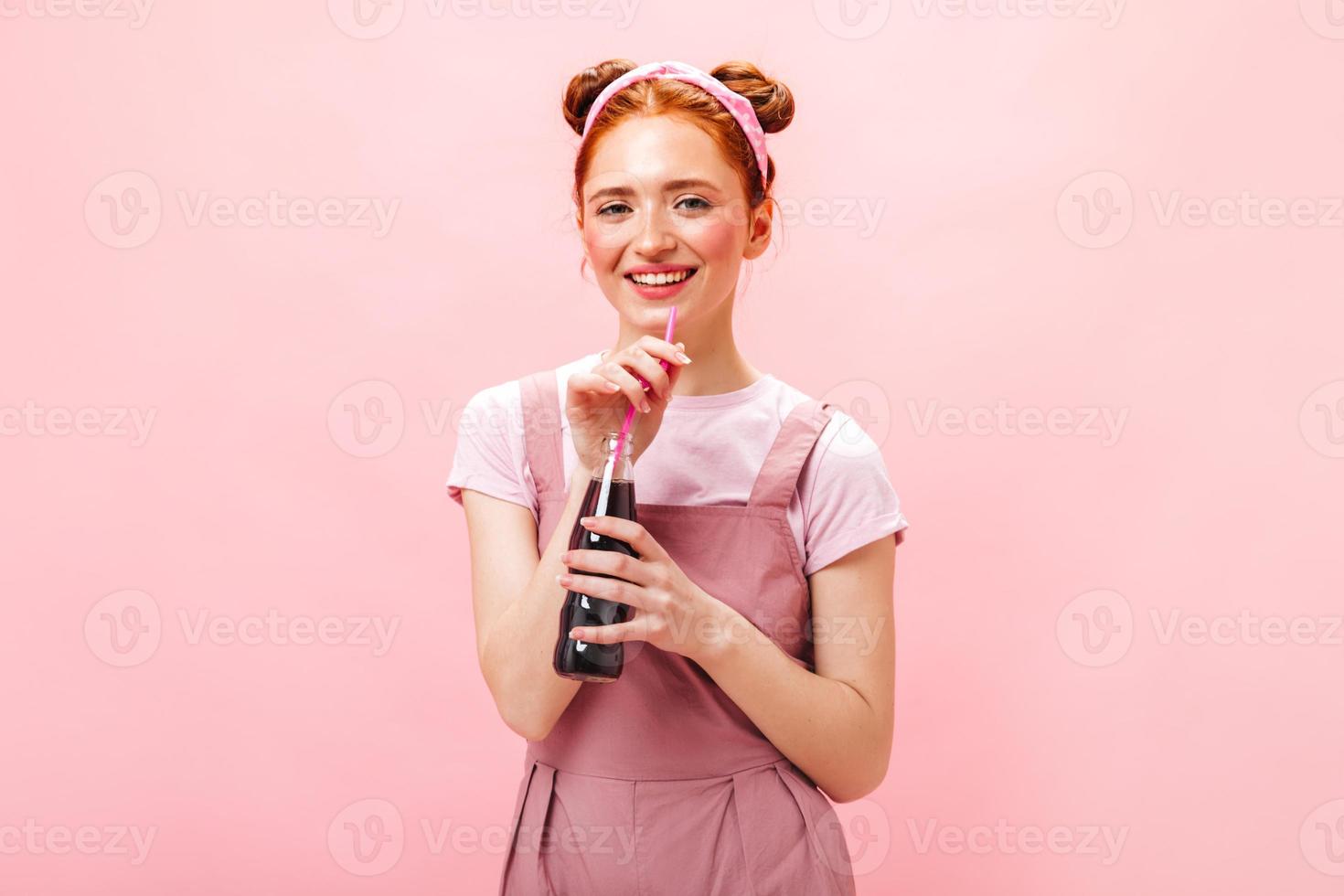 jovem alegre de vestido rosa mostra sinal de paz, sorri e segura garrafa de refrigerante no fundo rosa foto