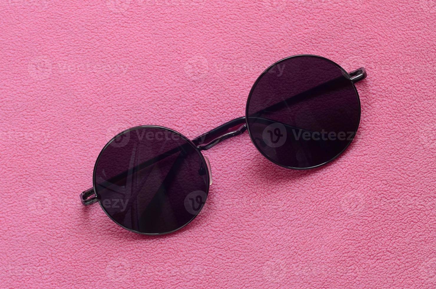 elegantes óculos de sol pretos com óculos redondos encontram-se em um cobertor feito de tecido de lã rosa claro macio e fofo. imagem de fundo elegante em cores femininas foto