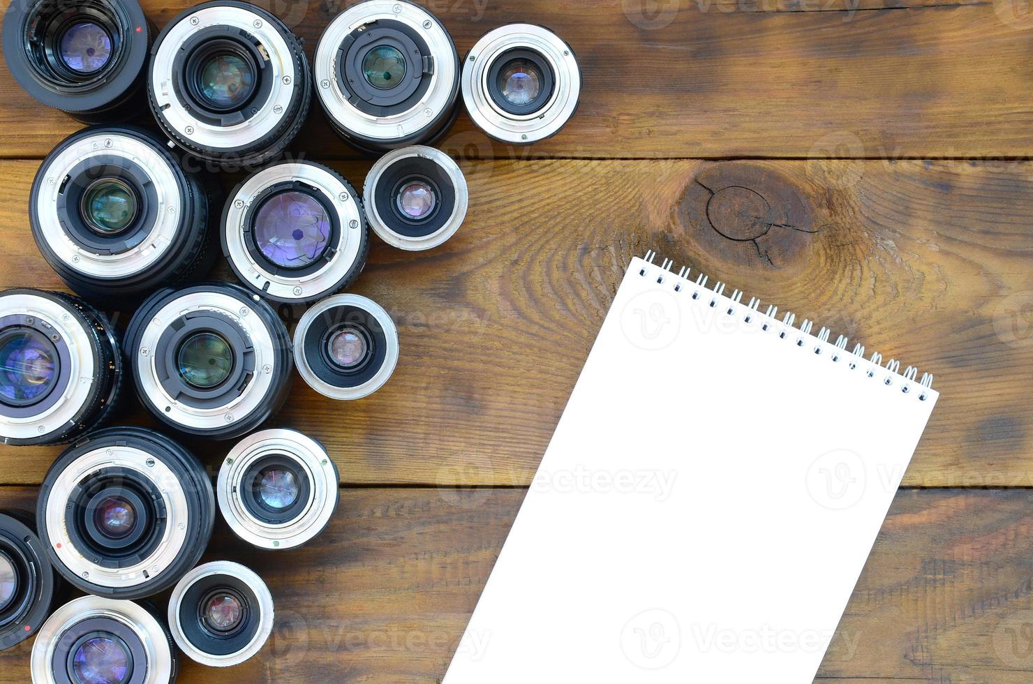 várias lentes fotográficas e caderno branco estão sobre um fundo de madeira marrom. espaço para texto foto