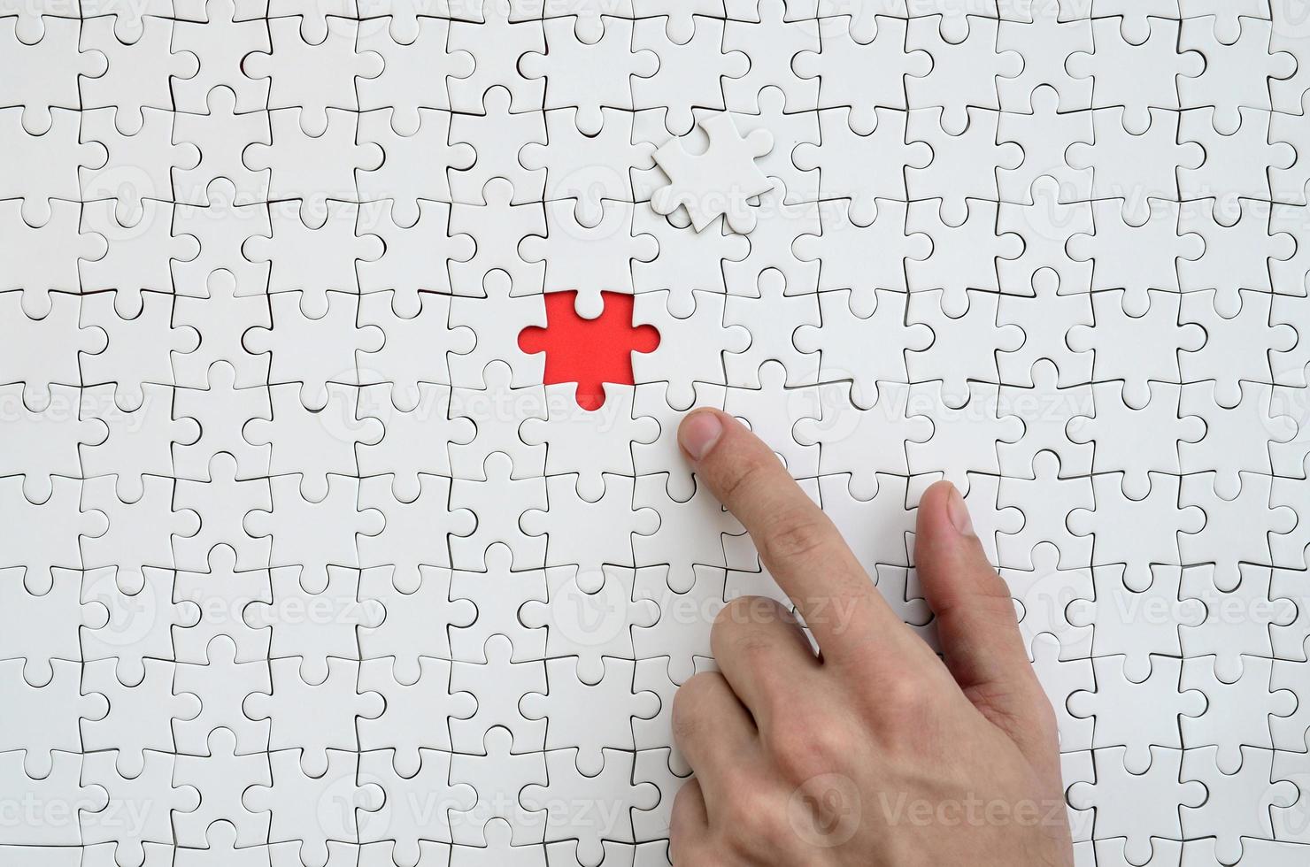 a textura de um quebra-cabeça branco no estado montado com um elemento faltando, formando um espaço vermelho, apontado pelo dedo da mão masculina foto