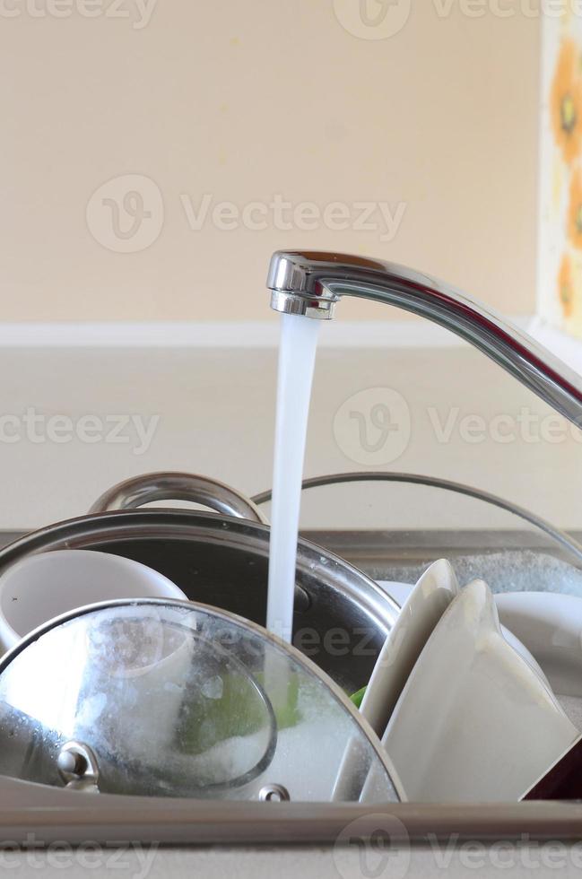 pratos sujos e utensílios de cozinha não lavados estão em água de espuma sob uma torneira de uma torneira de cozinha foto