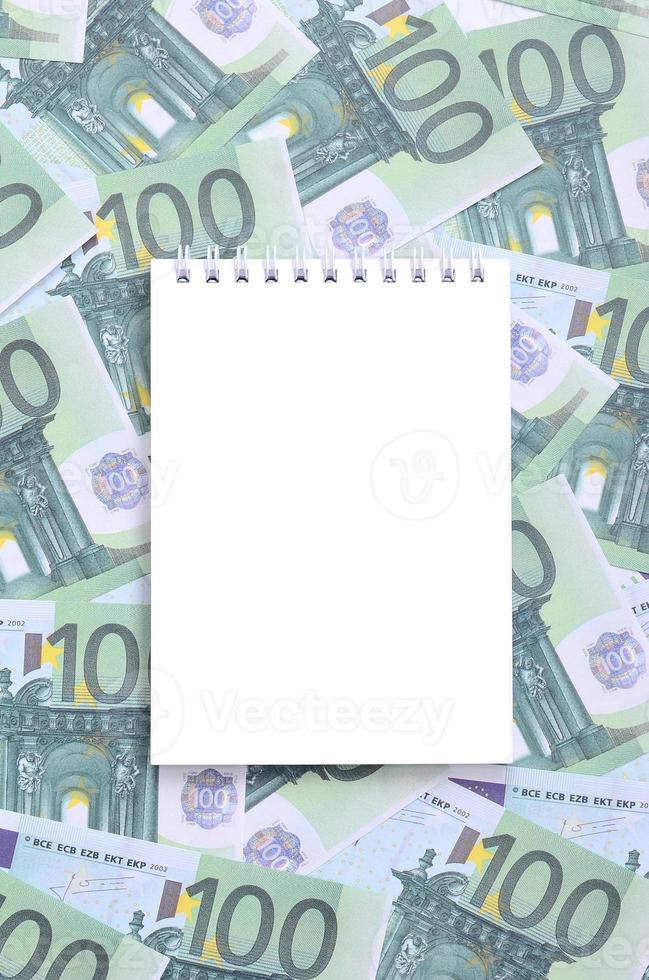caderno branco com páginas limpas sobre um conjunto de denominações monetárias verdes de 100 euros. muito dinheiro forma uma pilha infinita foto