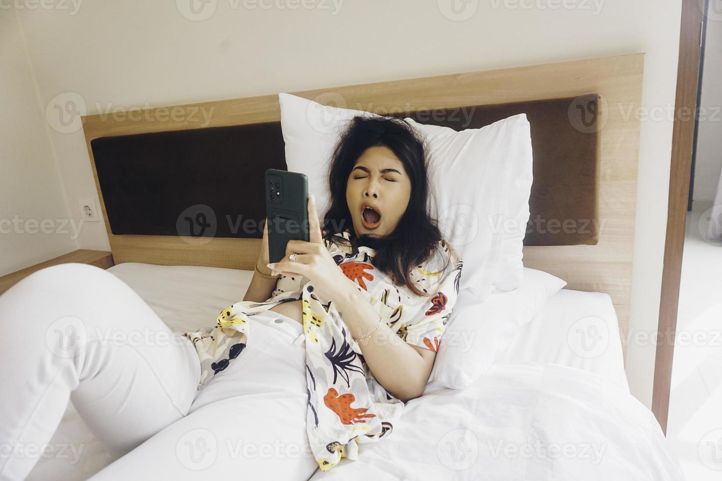 uma jovem com sono bocejando enquanto segura o telefone na cama. foto