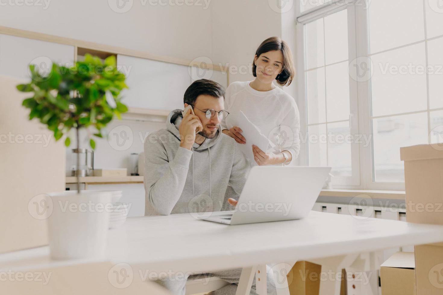 foto de modelo masculino pesquisa informações no computador portátil, procura um novo apartamento para alugar, liga para o cliente via celular, mulher fica perto com documentos em papel, verifica contas domésticas, pose na cozinha