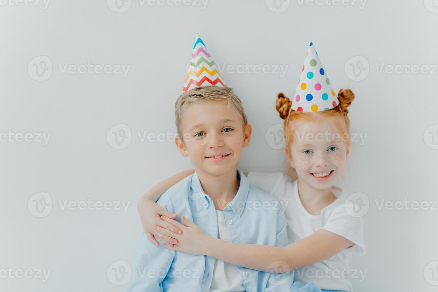 linda garota ruiva abraça com amor seu irmão mais velho, parabeniza pelo aniversário, usa chapéus de festa de cone, tem bom humor na festa, isolado sobre fundo branco, tem relacionamento amigável foto