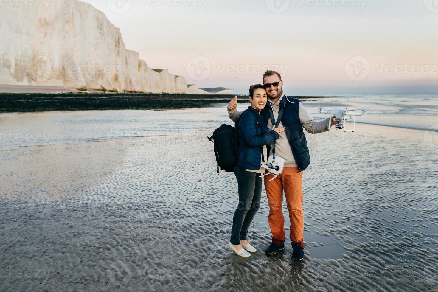 retrato ao ar livre de um lindo casal afetuoso, abraçando-se, opere com drone pelo controle de remont à beira-mar. jovens turistas femininos e masculinos navegam em quadricóptero. turismo e tecnologia foto