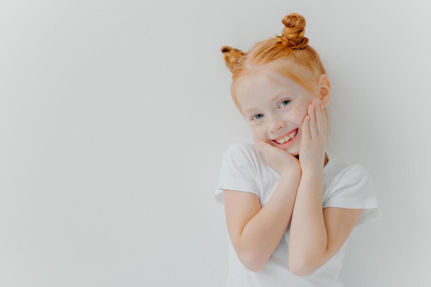 menina bonita tímida tem dois coques de cabelo, toca as bochechas suavemente, sorri alegremente, tem sardas, ouve algo positivo dos pais, veste camiseta branca, fica de pé, espaço em branco deixado para sua informação foto