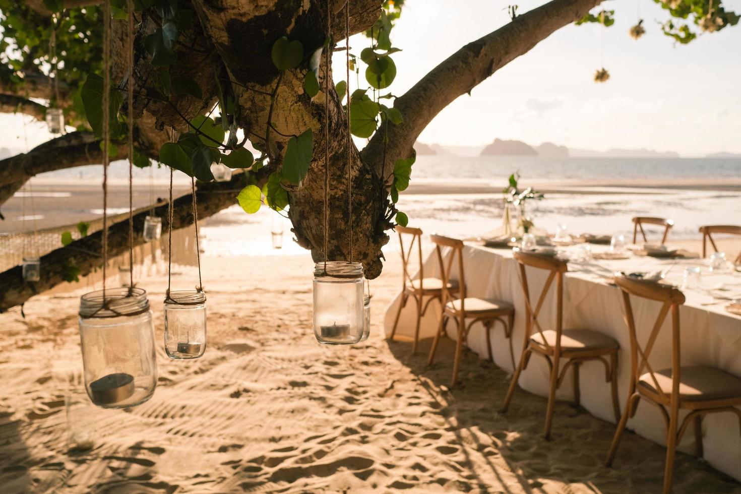 feche a vela do frasco pendurada na árvore com a configuração do jantar de casamento de mesa longa na praia na tailândia à noite. conceito de festa de casamento. decoração restaurante ao ar livre na praia. foto