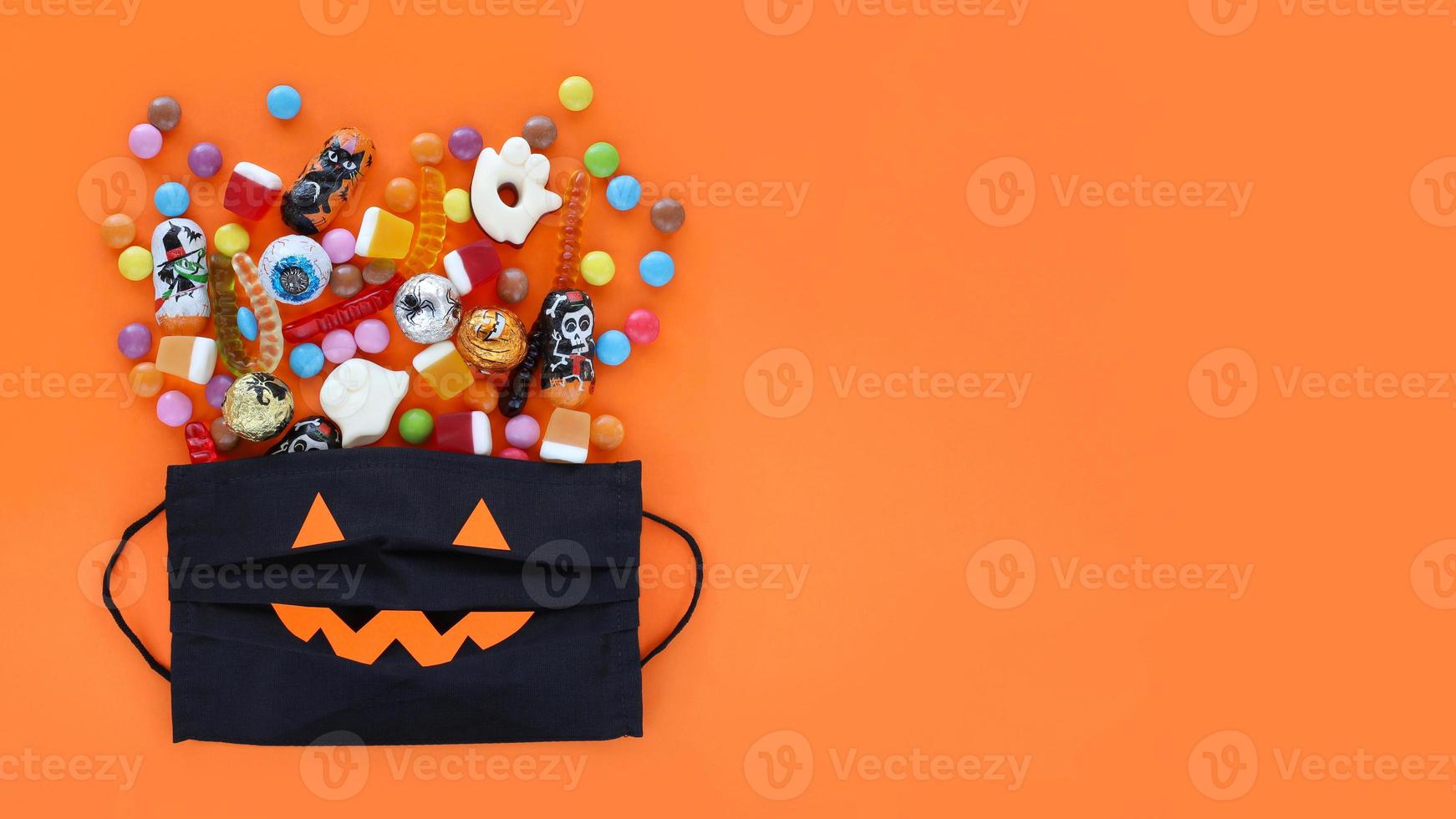 máscara médica preta como uma bolsa de presentes de abóbora sorridente cheia de doces tradicionais de halloween variados derramados. fundo de banner laranja com espaço de cópia. foto