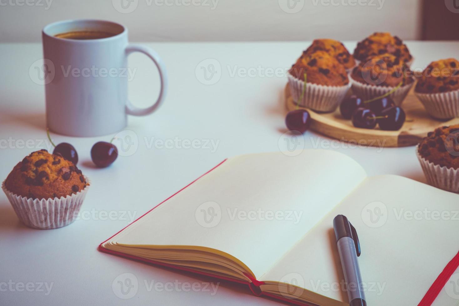 um caderno vazio com uma capa vermelha sobre uma mesa ao lado de uma caneta, uma xícara de café, muffins de chocolate e algumas cerejas foto