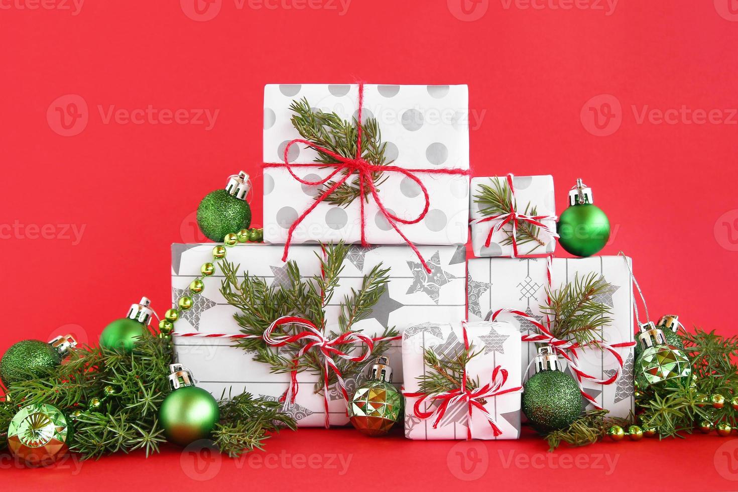 caixas de presente de natal embrulhadas em papel branco-cinza com fitas vermelho-brancas sobre fundo vermelho, decoradas com galhos de abeto e decorações de natal verdes brilhantes. conceito de natal e ano novo. foto