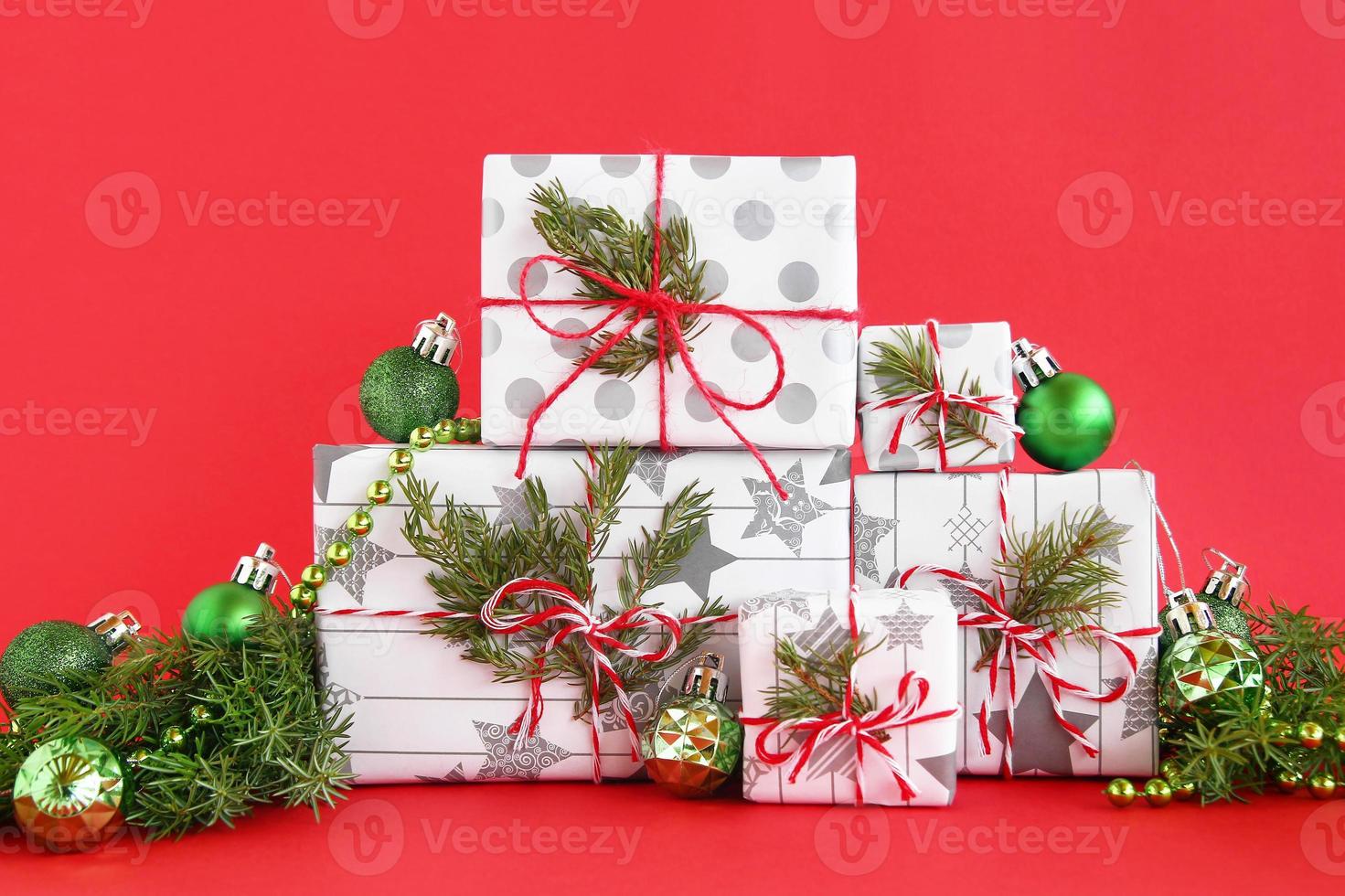 caixas de presente de natal embrulhadas em papel branco-cinza com fitas vermelho-brancas sobre fundo vermelho, decoradas com galhos de abeto e decorações de natal verdes brilhantes. conceito de natal e ano novo. foto