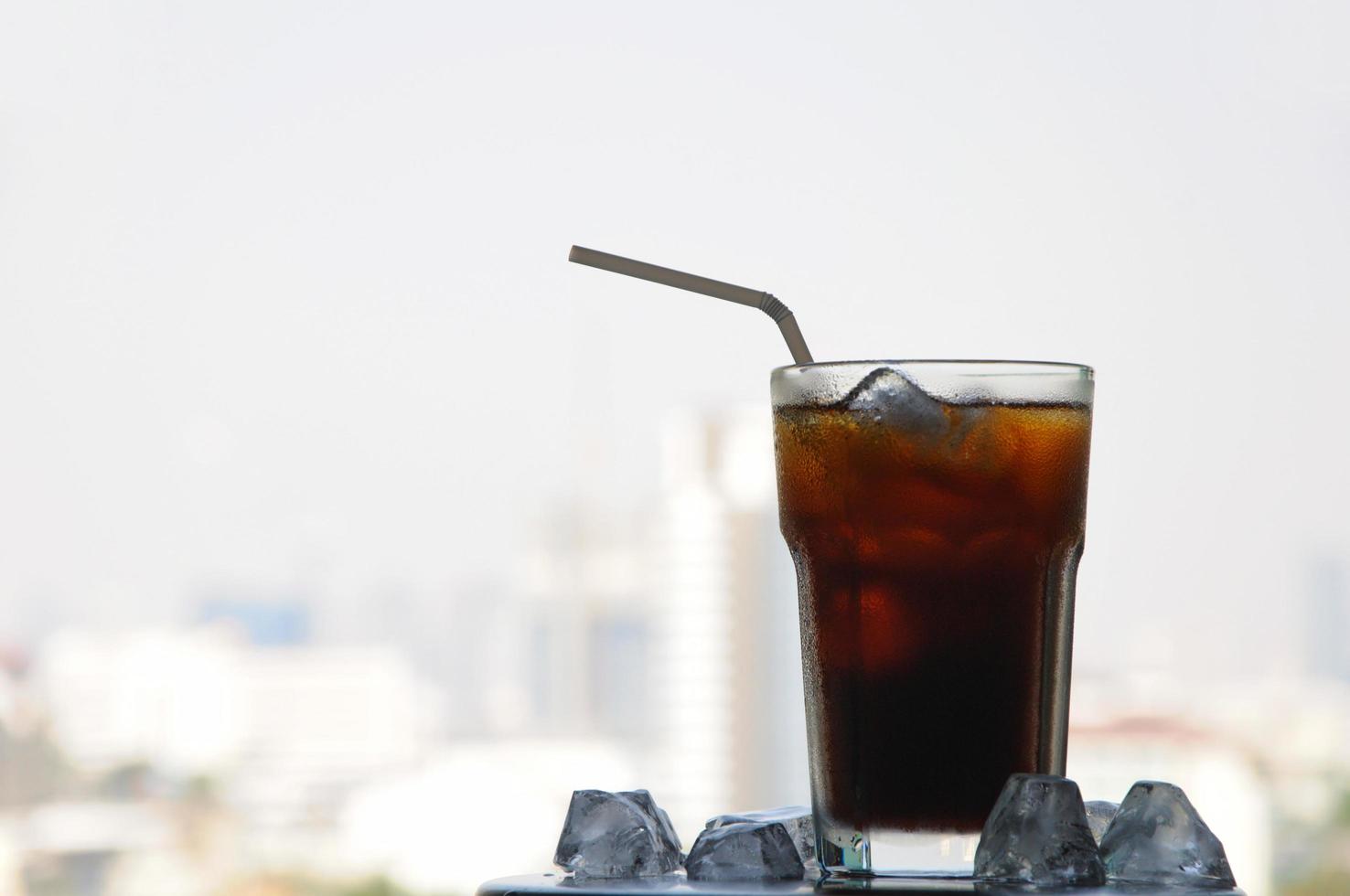 um copo de café preto gelado na mesa com cubo de gelo para aproveitar o clima quente no verão com o fundo da vista da cidade foto