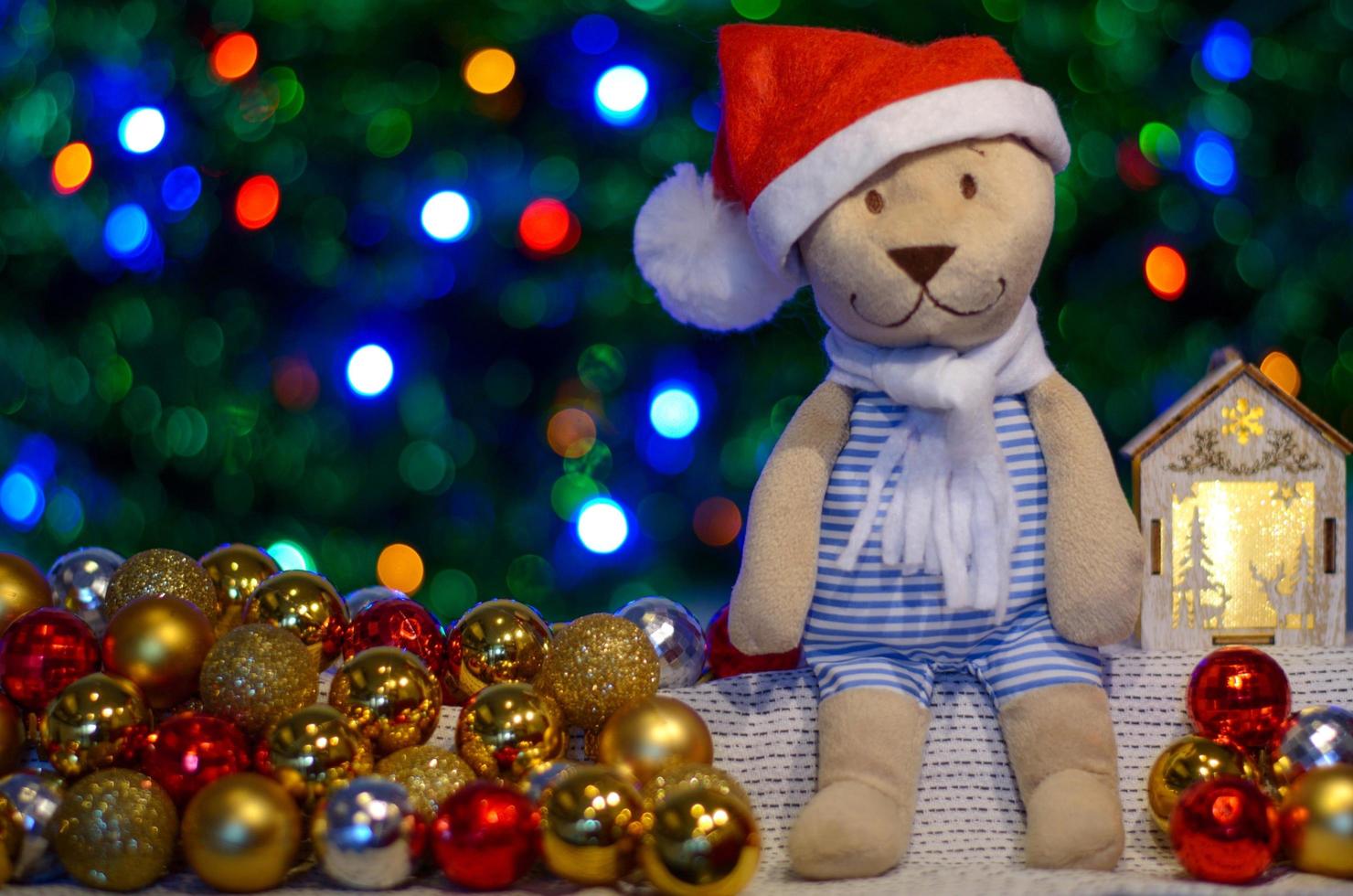 ursinho de pelúcia de papai noel sentado na mesa com bugiganga e árvore de natal e fundo de luzes coloridas bokeh. foto