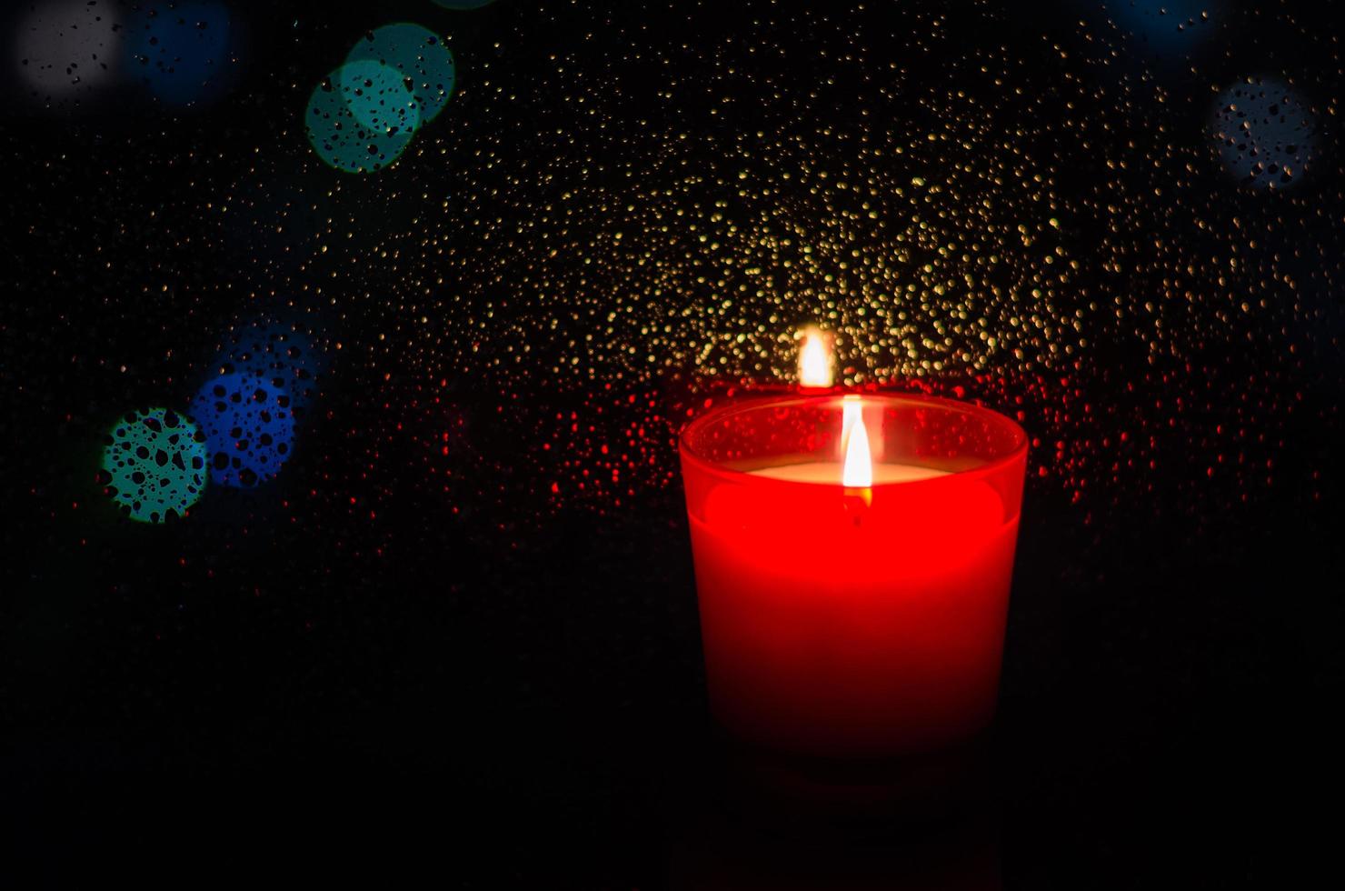 vela acesa em vidro vermelho coloca ao lado da janela com gota de chuva em bokeh luz e fundo escuro. foto